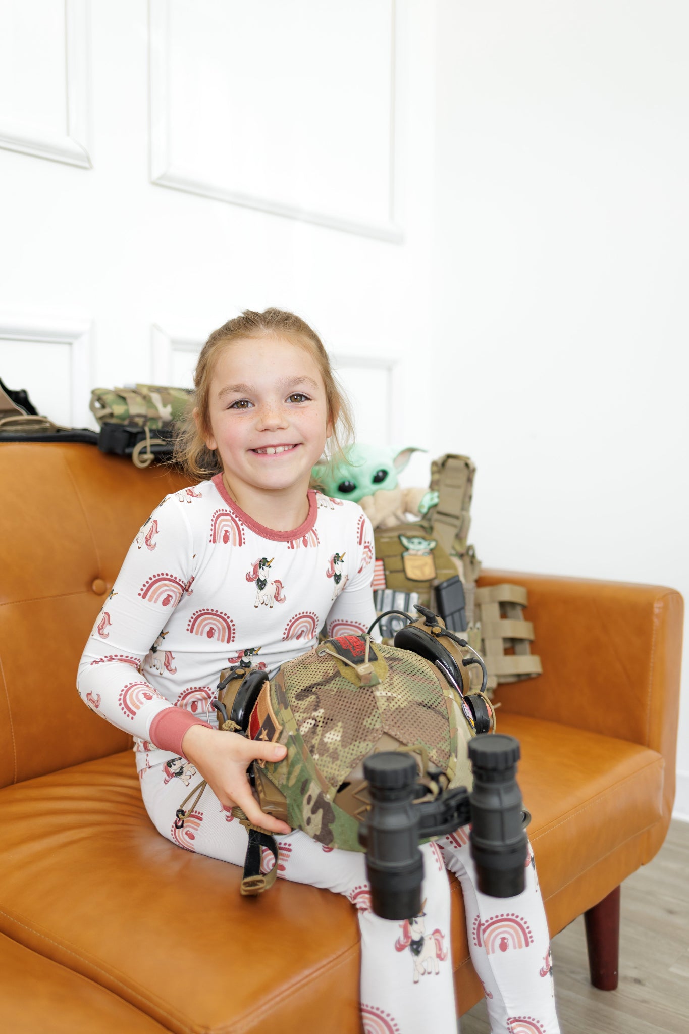 Child in pajamas holding a toy gun and helmet on a brown couch with toys in the background.