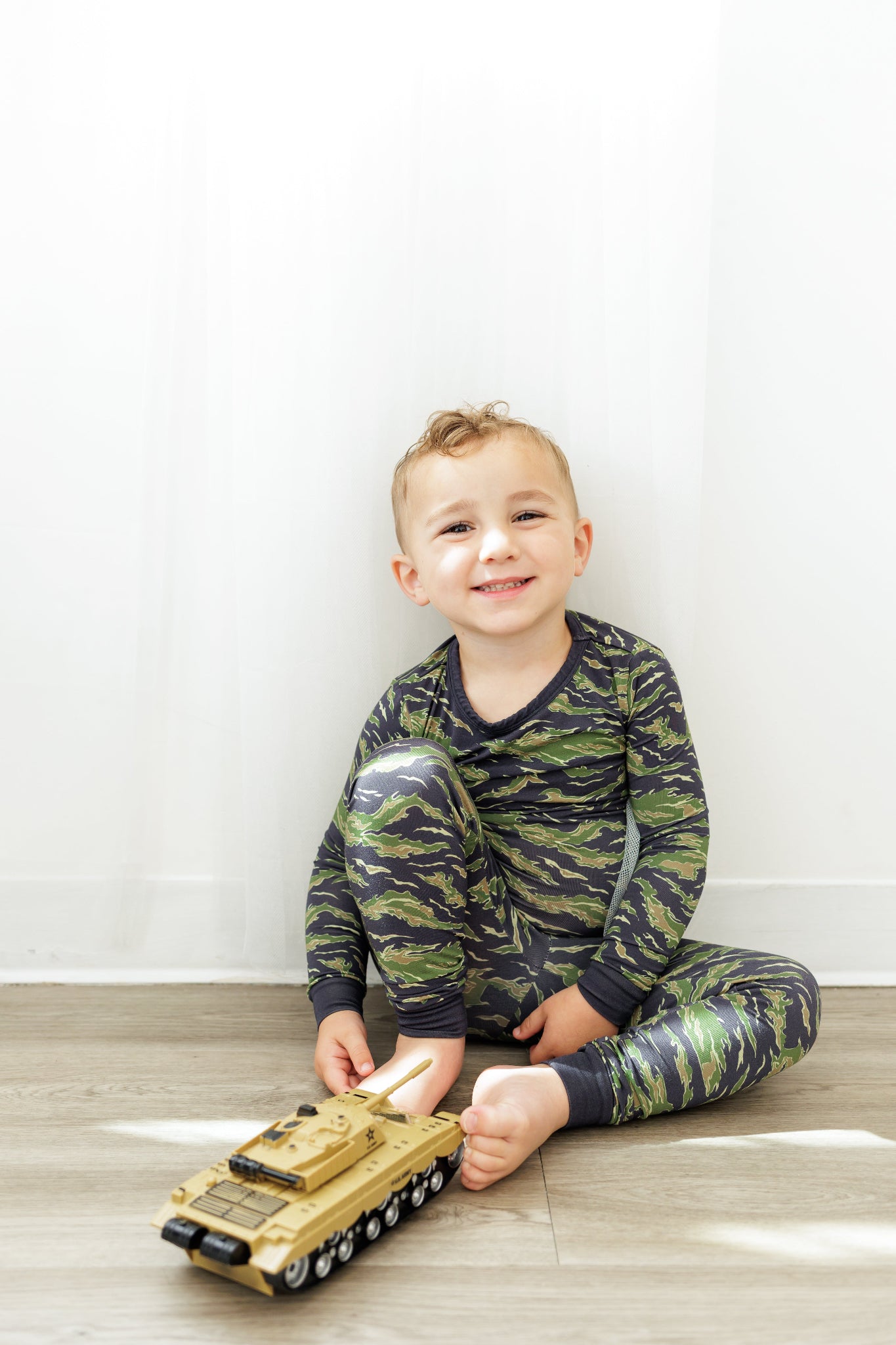 Child wearing camouflage pajamas playing with a toy tank on a wooden floor.