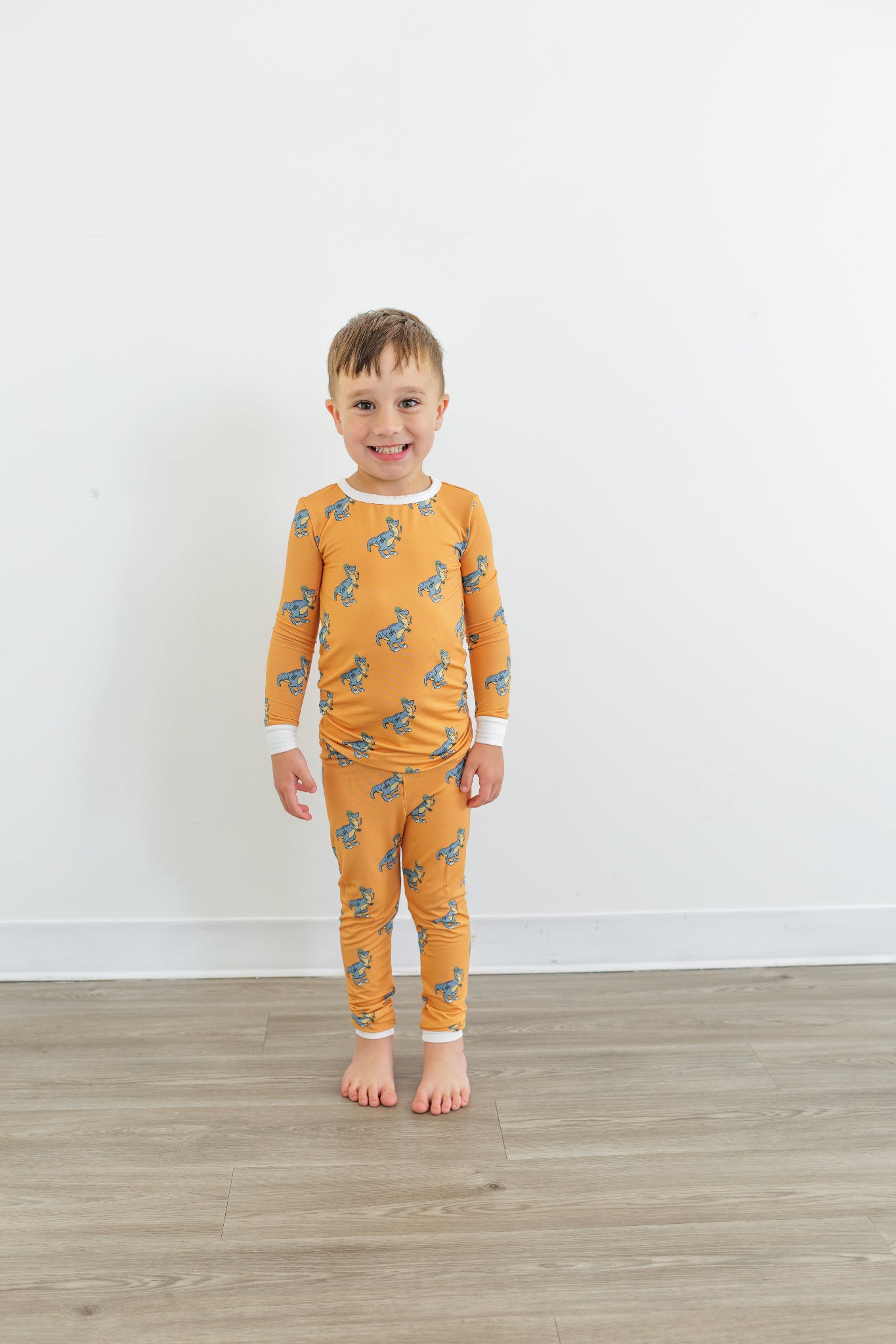 Child wearing orange dinosaur-patterned pajamas standing on a wooden floor with a white wall background