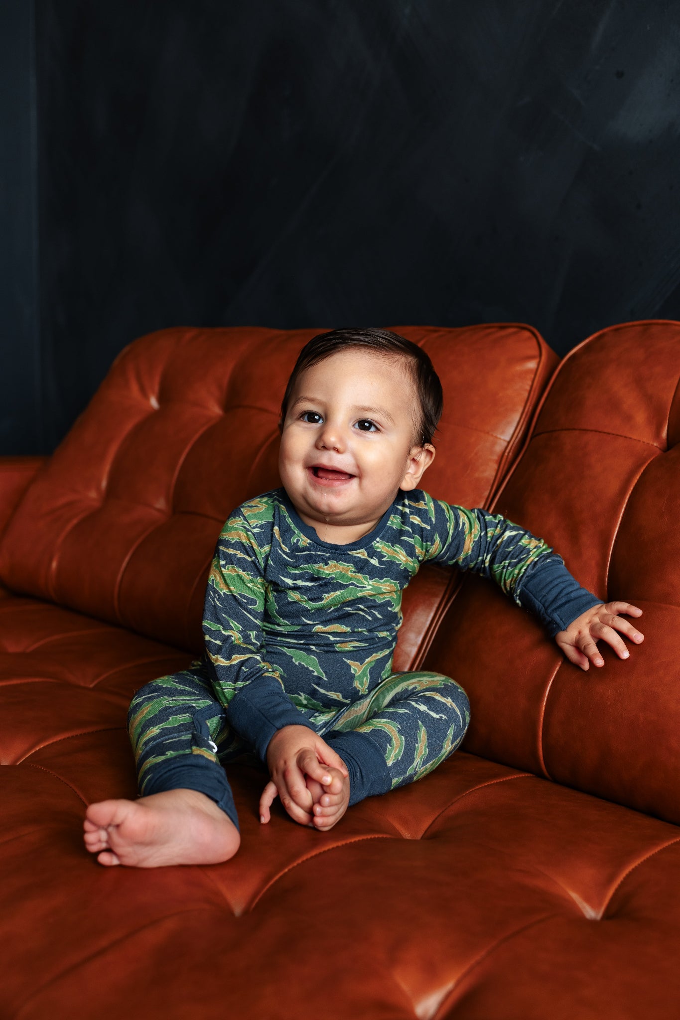 Child wearing a blue and green patterned onesie sitting on a leather couch against a dark background