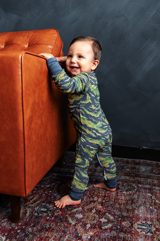 Child in a camouflage onesie standing next to an orange couch against a dark wall.