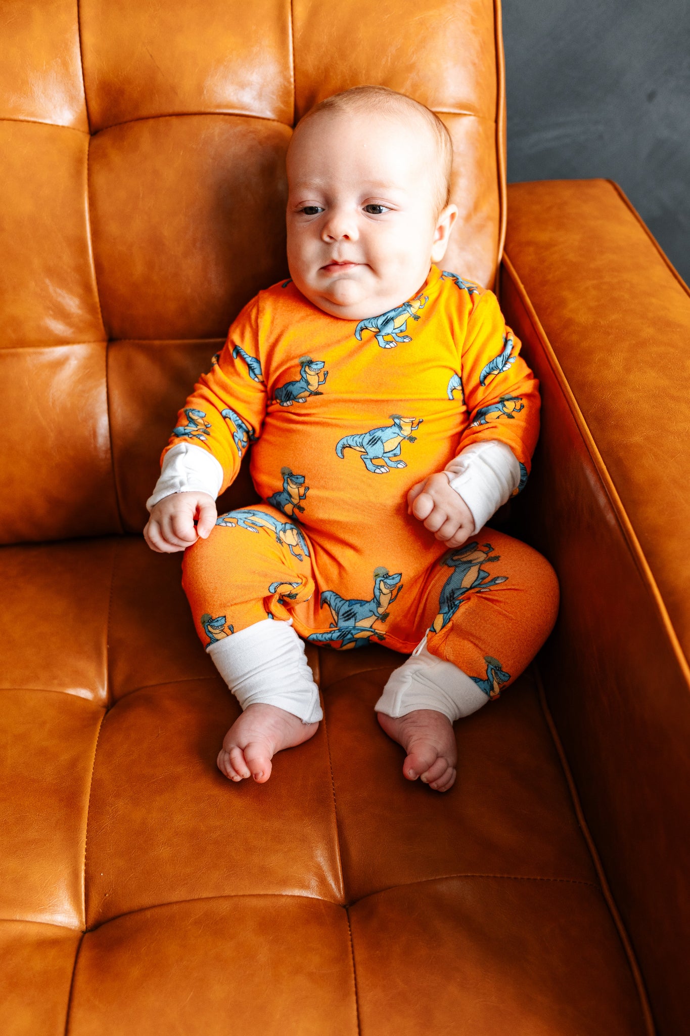 Baby in an orange outfit with dinosaur patterns sitting on a brown leather chair.