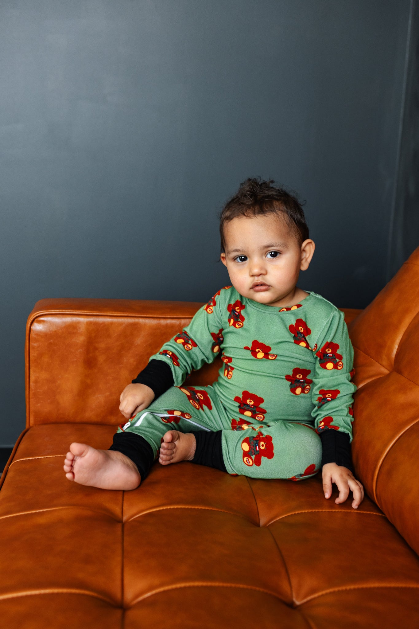 Child wearing a green outfit with red floral patterns sitting on a brown leather couch against a dark gray wall.