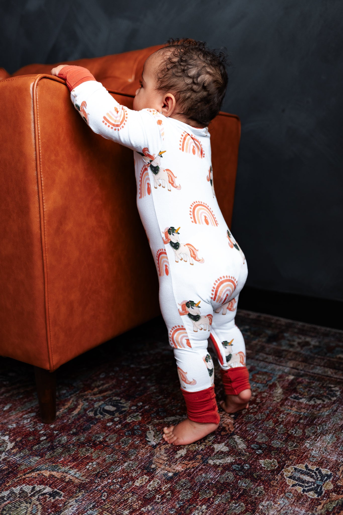 Child in a patterned onesie standing next to an orange chair on a textured rug.