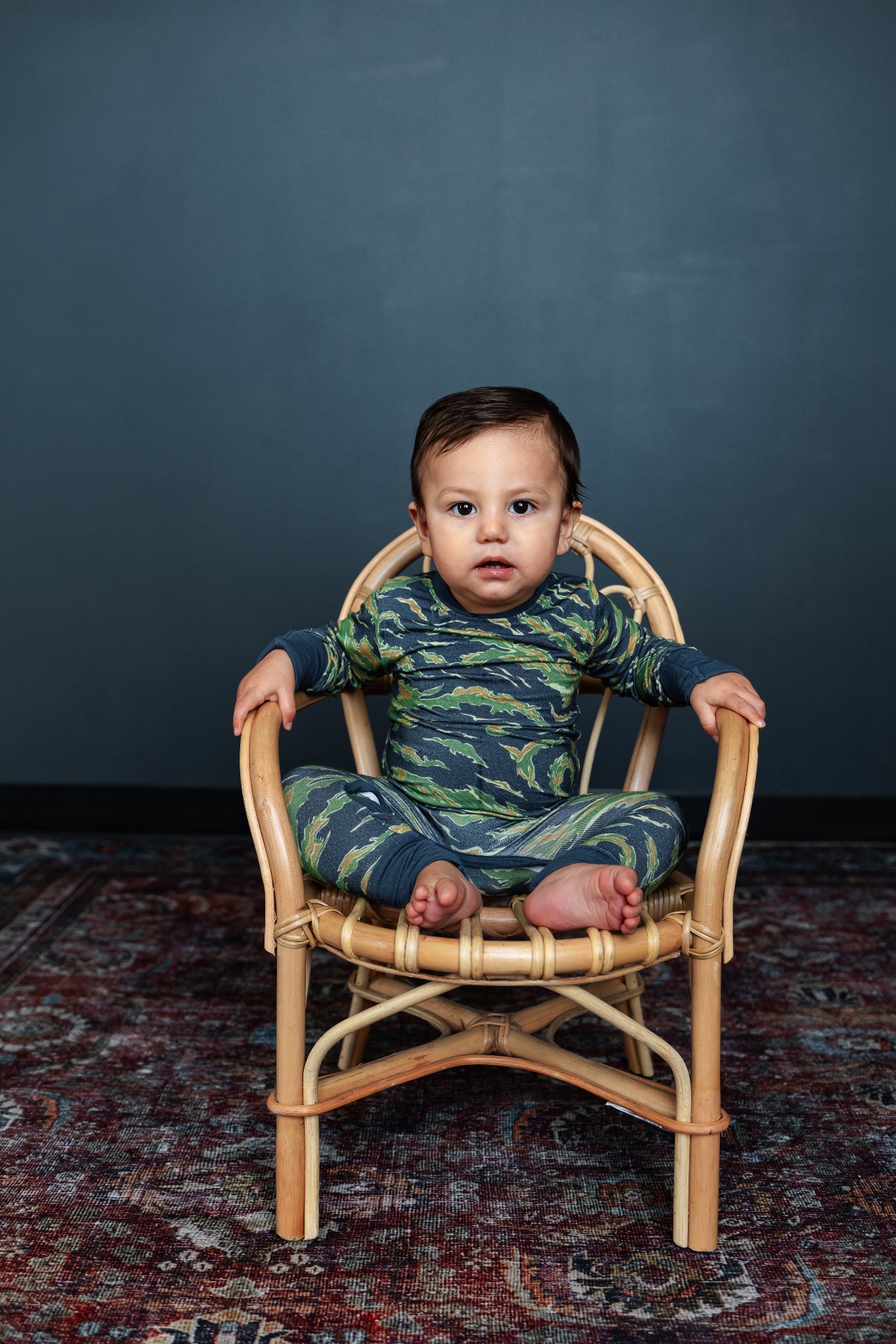 Baby in a camouflage outfit sitting on a small wooden chair against a dark background