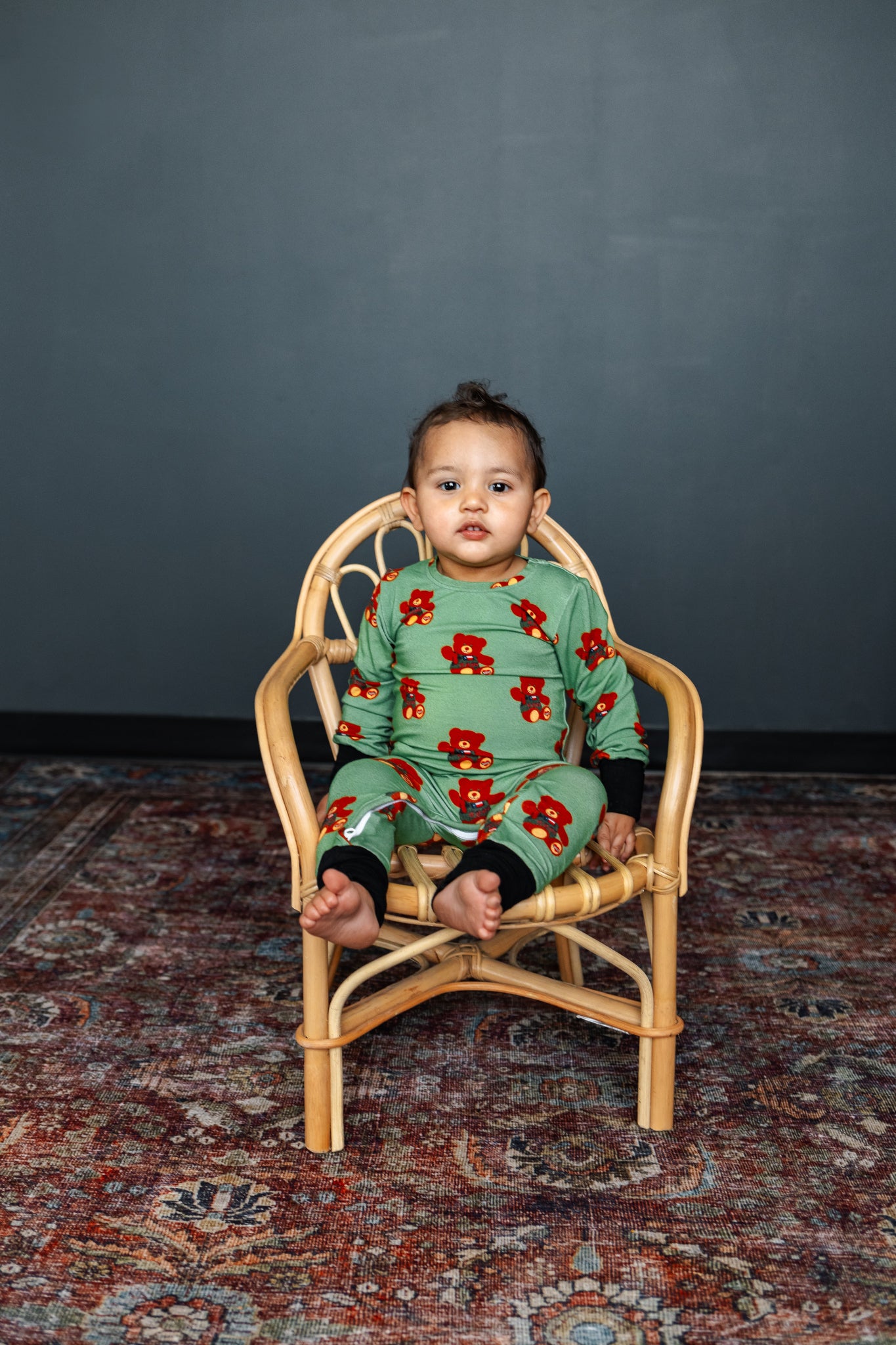 Child wearing a green floral onesie sitting on a small wooden chair against a dark gray wall.