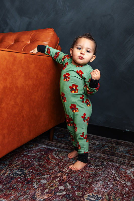 Child wearing a green onesie with red floral patterns standing next to an orange couch against a dark wall.