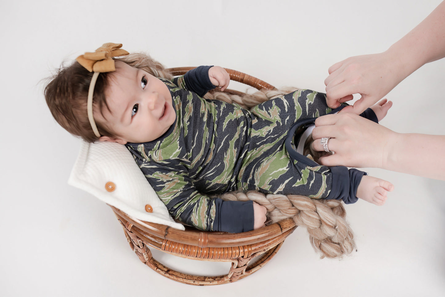 Smiling baby lying in a wicker basket wearing a green and brown camo romper with navy trim, as an adult zips open the inseam for an easy diaper change. Baby wears a mustard-colored bow headband.