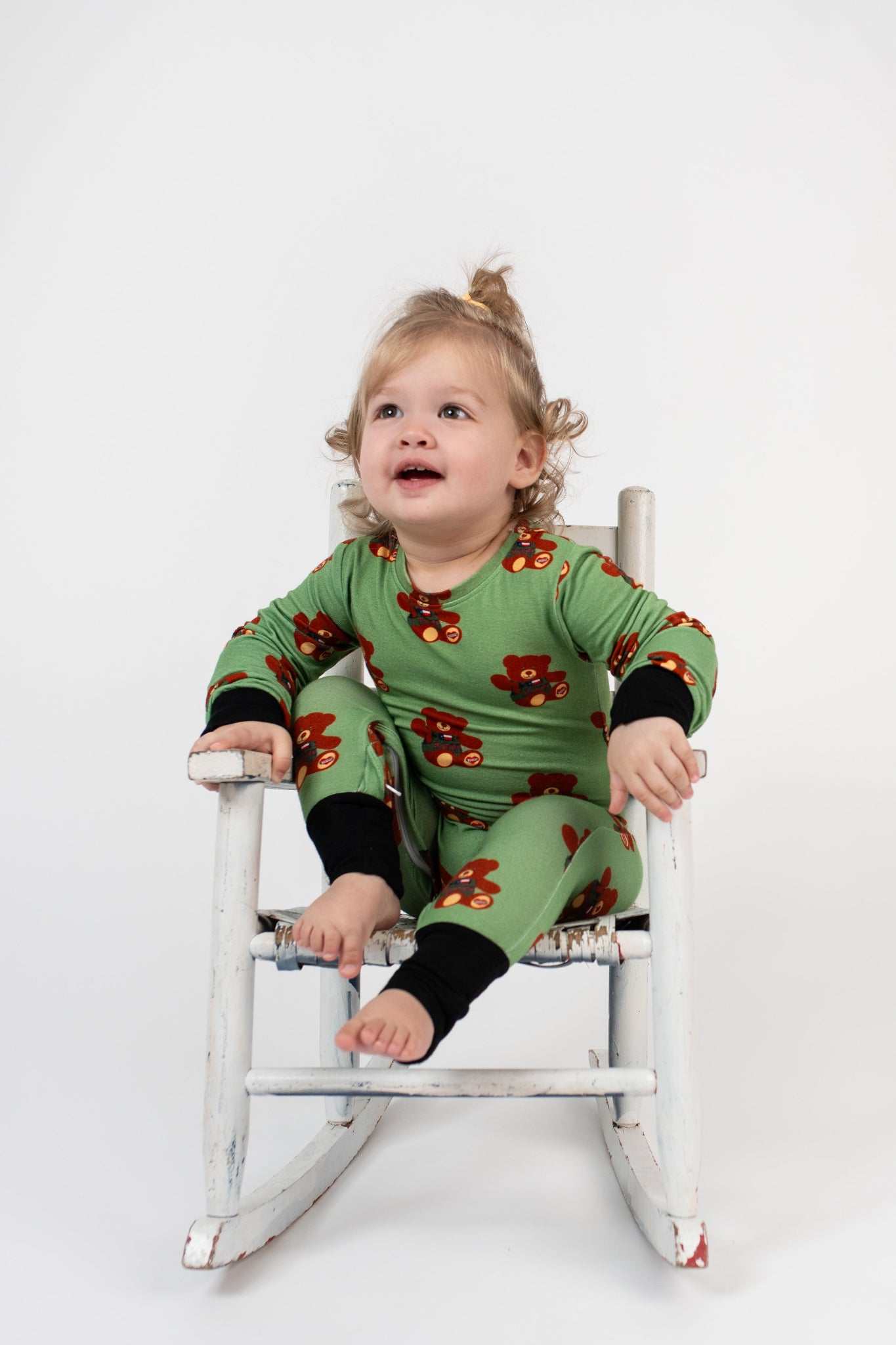 Toddler sitting in a vintage white rocking chair, wearing a green romper with black cuffs and a repeating tactical teddy bear print from 3rd Coast Limited.