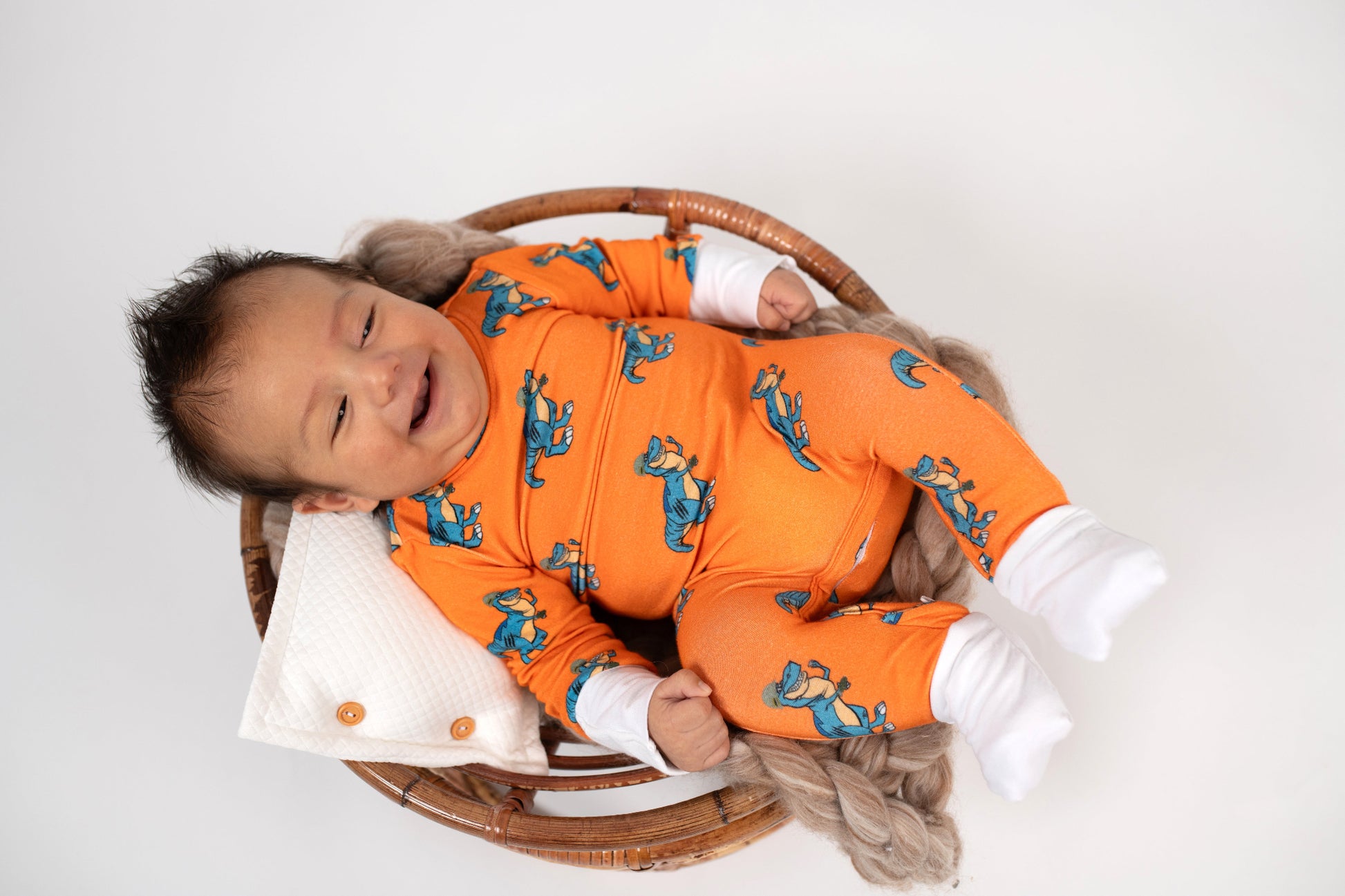Smiling baby lying in a round wicker basket, wearing an orange romper with a blue T. rex print and white cuffs from 3rd Coast Limited.