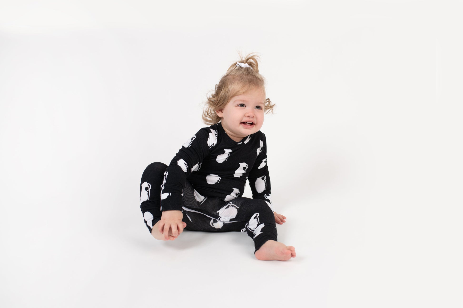 Smiling toddler sitting on a white backdrop, wearing a black long-sleeve romper with a white grenade pattern from 3rd Coast Limited.