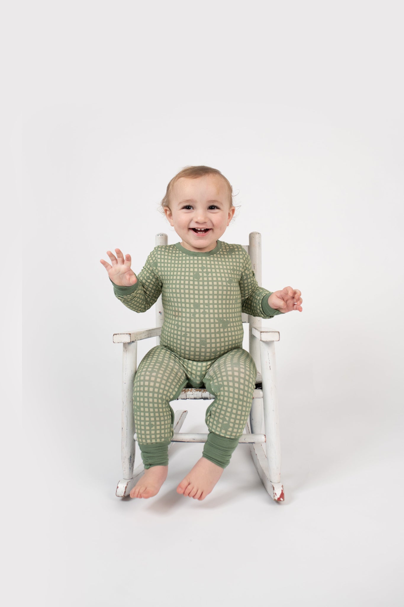 Smiling toddler sitting in a vintage white rocking chair, wearing a sage green romper with a tan grid pattern and matching green cuffs from 3rd Coast Limited.
