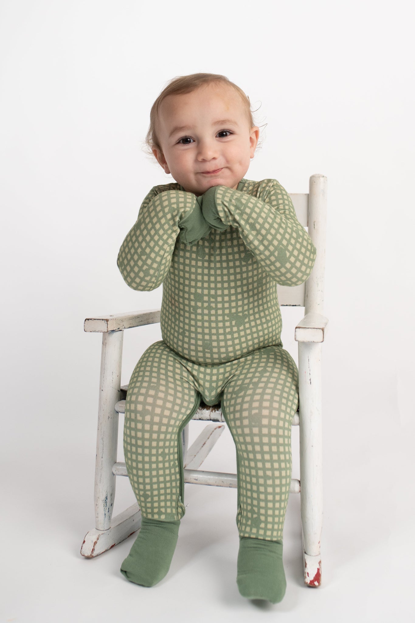 Smiling toddler sitting in a vintage white rocking chair, wearing a sage green romper with tan grid pattern, fold-over hand and foot cuffs covering hands and feet, from 3rd Coast Limited.