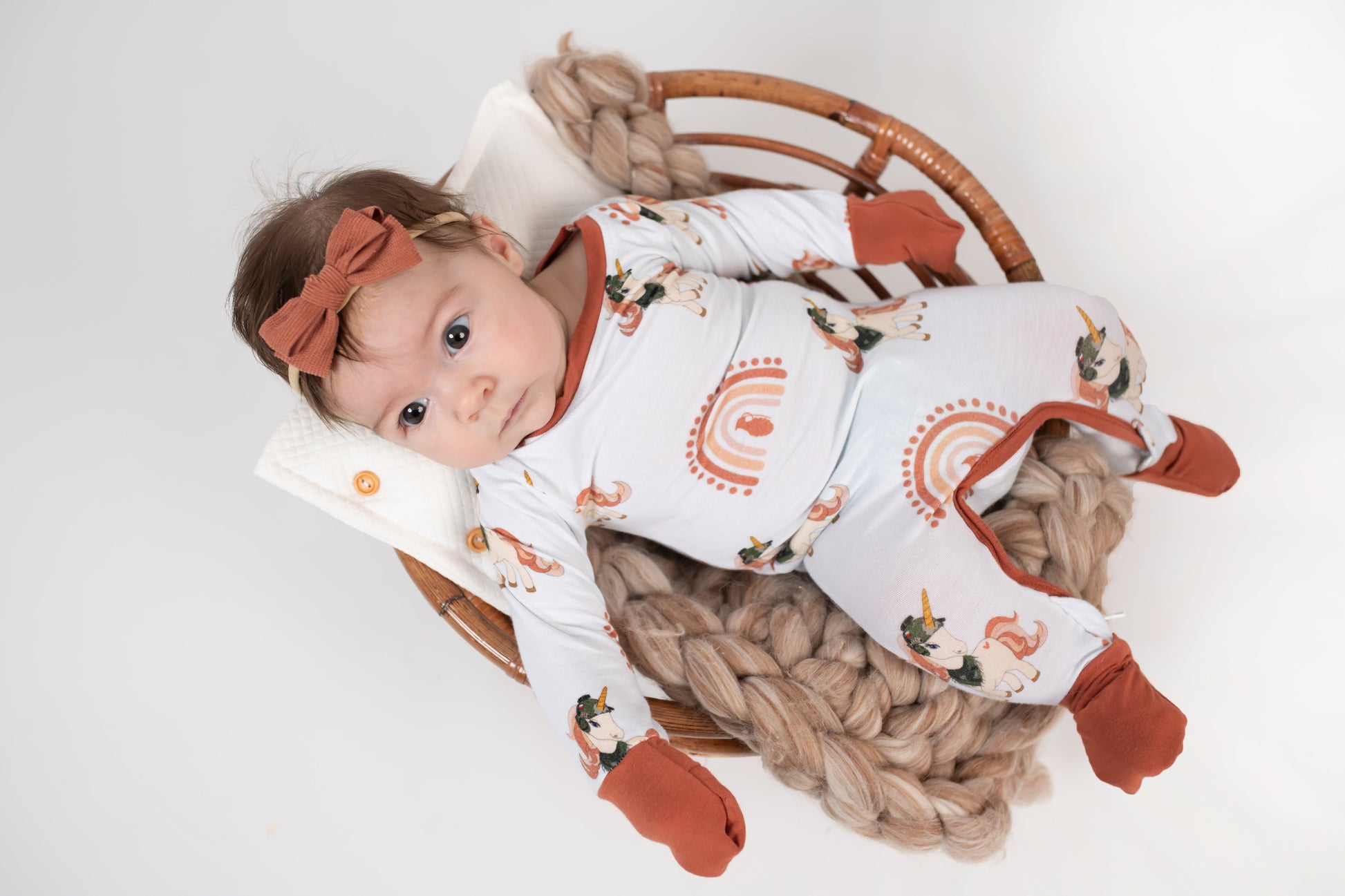 Baby lying in a round wicker basket on a chunky knit blanket, wearing a cream romper with rust cuffs, rainbow designs, and tactical unicorn print, paired with a rust-colored bow headband.