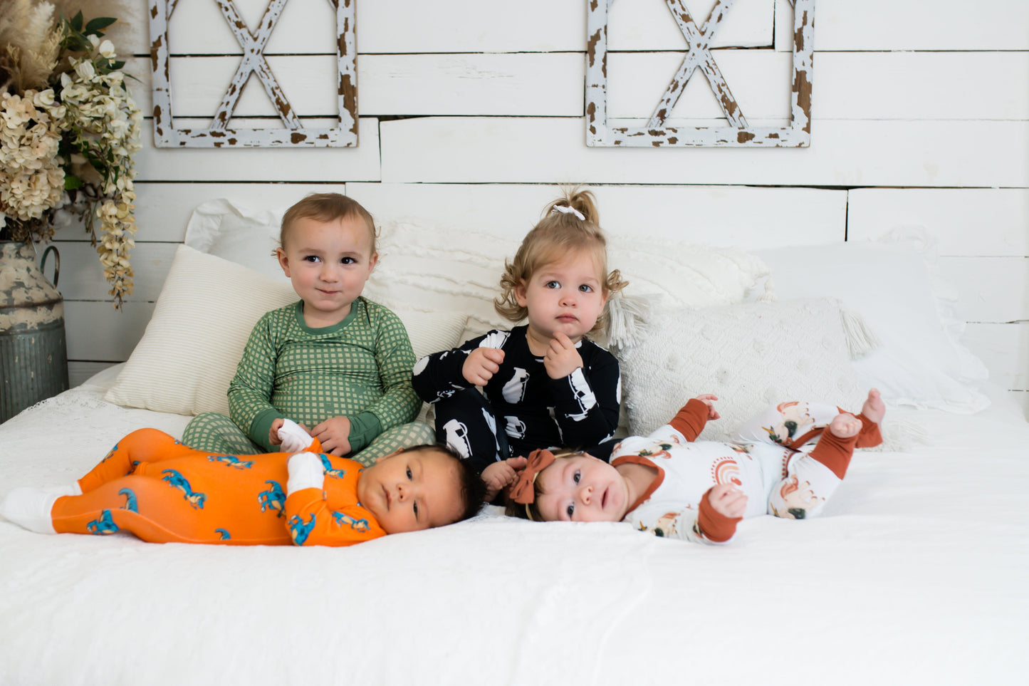 Four young children on a white bed in a farmhouse-style room, each wearing different printed pajamas—including green checkered, black with white grenades, bright orange with blue dinosaurs, and white with rust-colored rainbows and animals.
