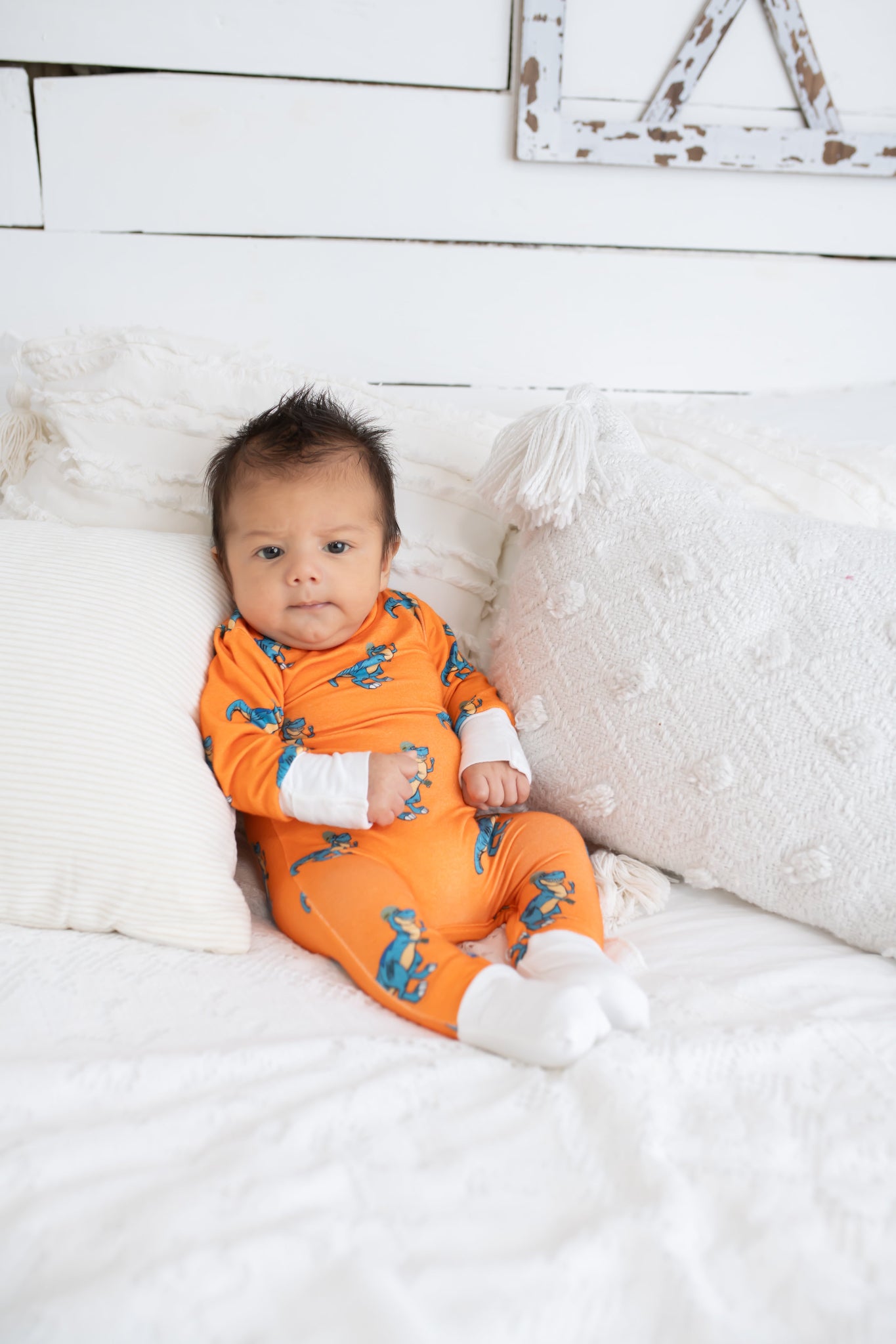 Baby sitting on a white bed with textured pillows, wearing an orange romper featuring a blue cartoon T. rex print and white cuffs from 3rd Coast Limited.