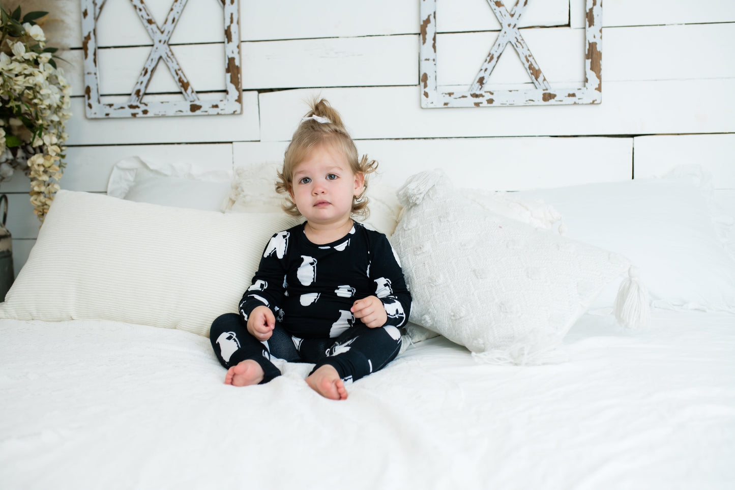 Toddler sitting on a white bed in a black romper with white grenade print, surrounded by textured white pillows and rustic farmhouse decor.