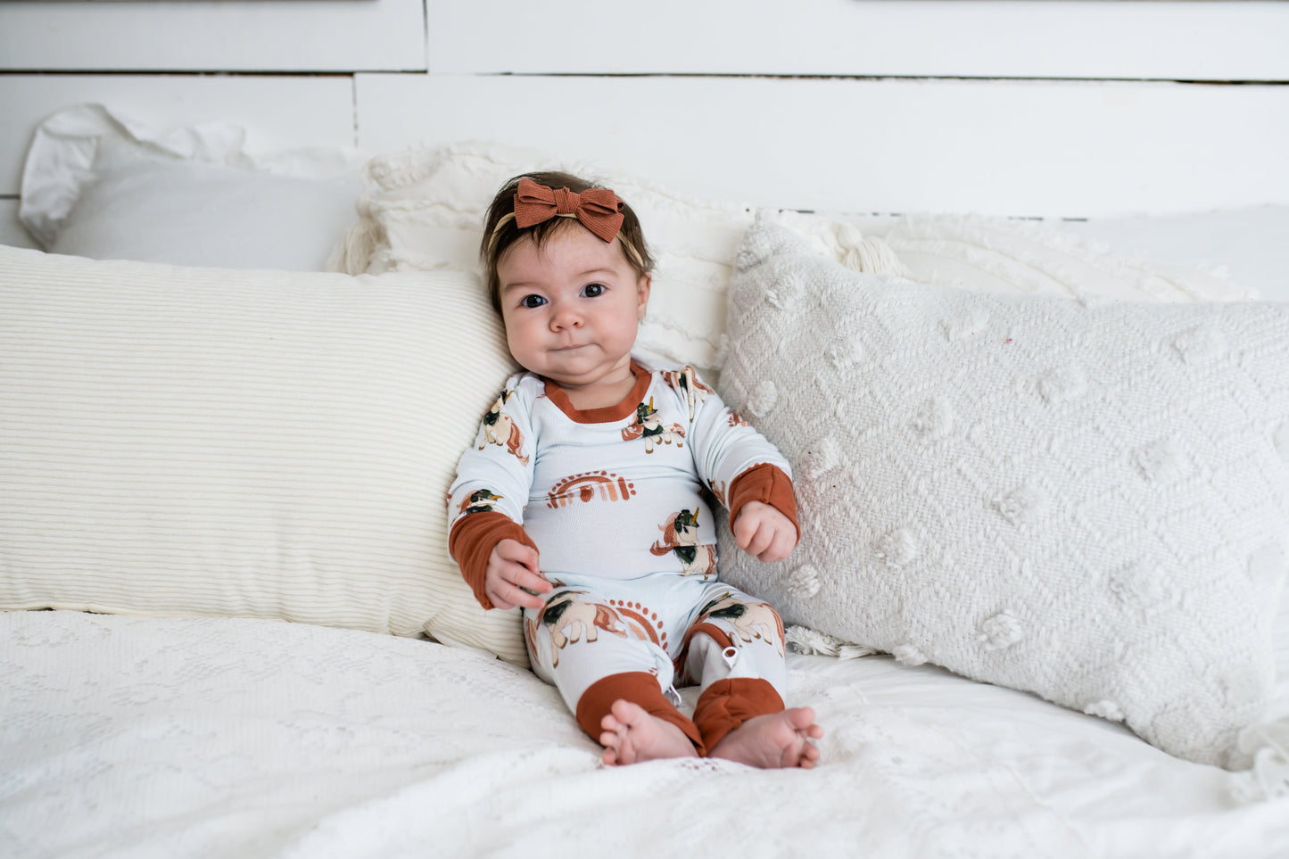 Baby sitting on a white bed with textured pillows, wearing a rust and cream-colored romper with rainbow and tactical print accents, and a matching rust bow headband.