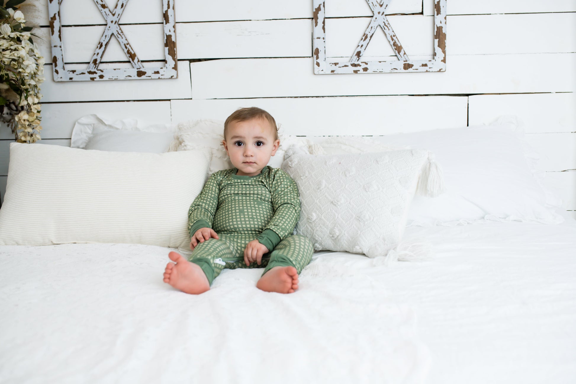 Toddler sitting on a white bed with textured pillows and farmhouse decor, wearing a sage green romper with a light tan grid pattern and matching green cuffs from 3rd Coast Limited.