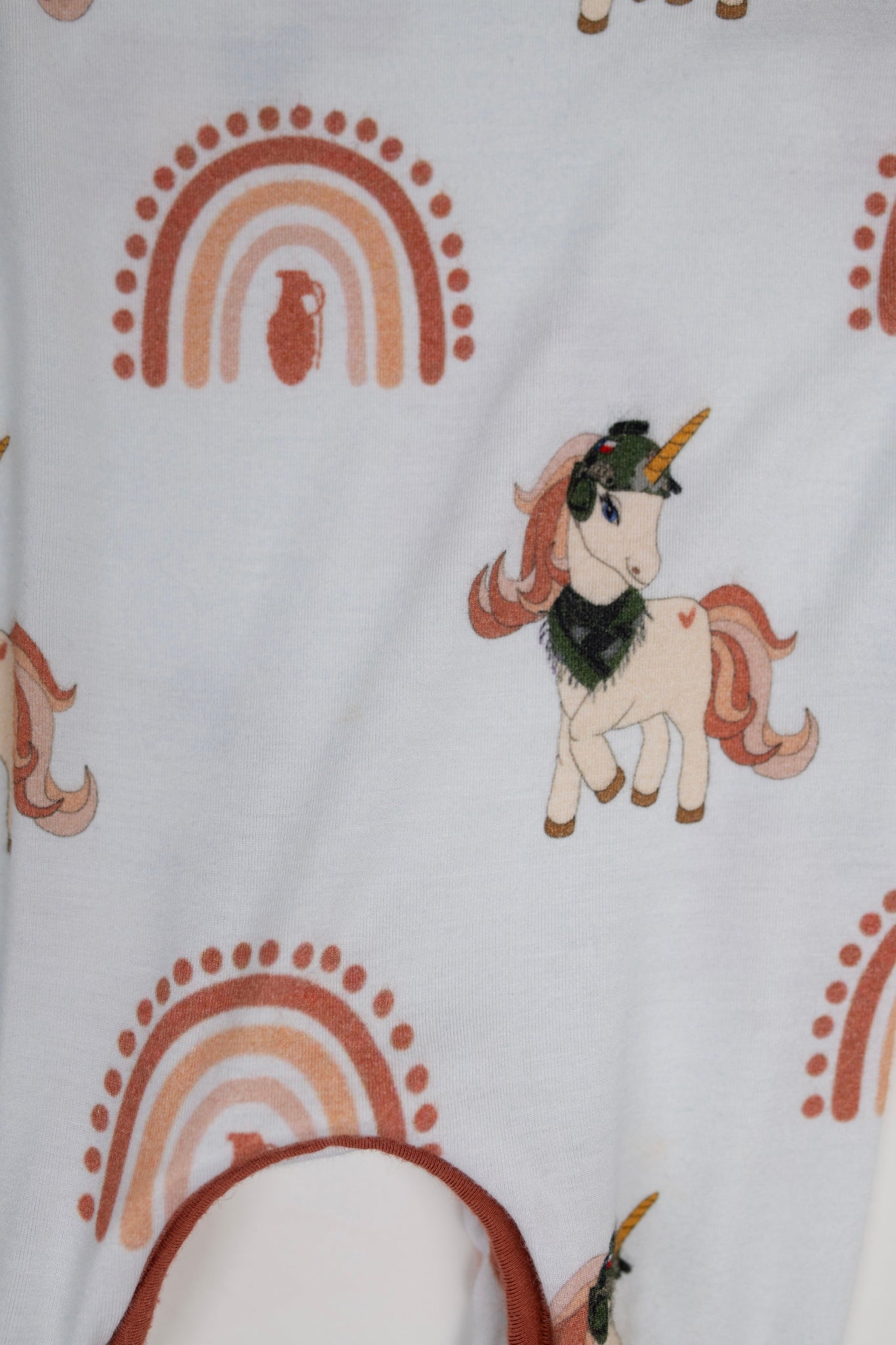 Close-up of cream baby romper fabric featuring a rust-colored rainbow with a grenade silhouette in the center and a tactical unicorn with a helmet and scarf, designed by 3rd Coast Limited.