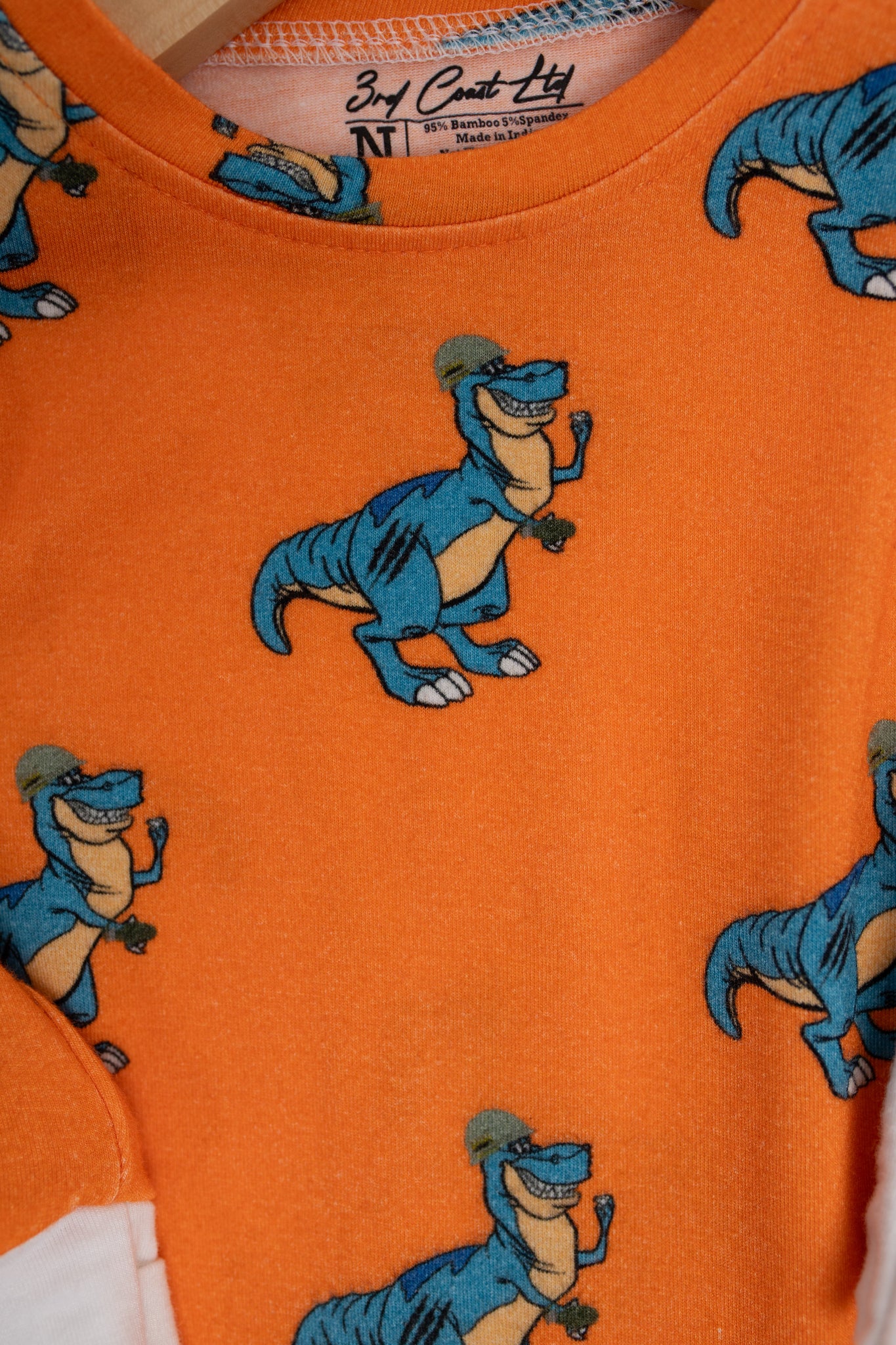 Close-up of an orange baby romper from 3rd Coast Limited, featuring a repeating print of a smiling blue T. rex wearing a military helmet and dog tags, flexing its arm.