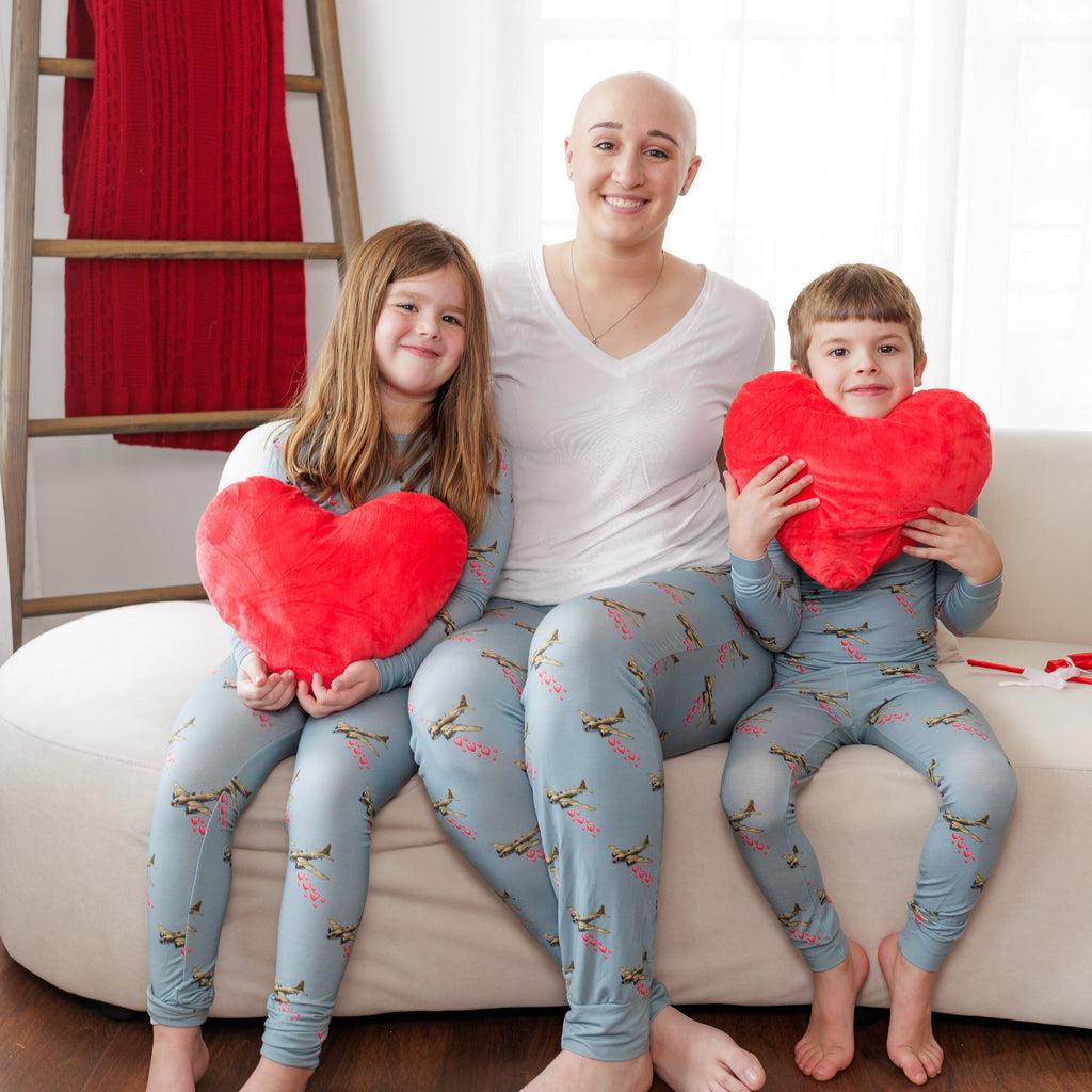 Woman and two children sitting on a couch holding red heart-shaped pillows in a home setting.