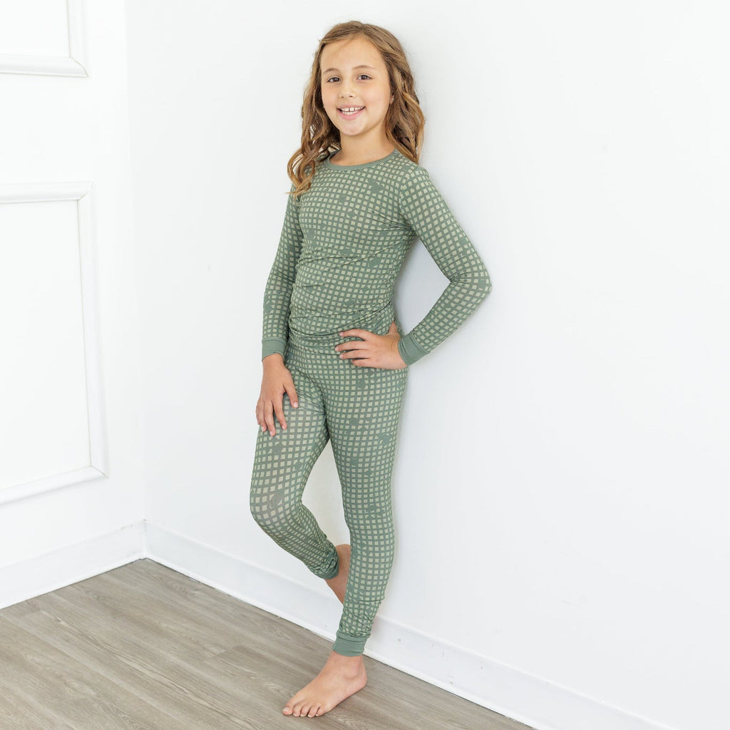 Young girl wearing green checkered pajamas standing in a room with white walls and wooden floor.