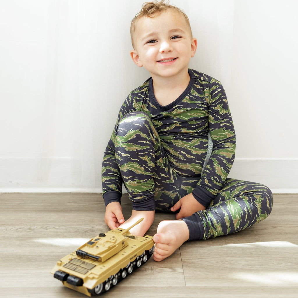 Child wearing camouflage pajamas playing with a toy tank on a wooden floor.