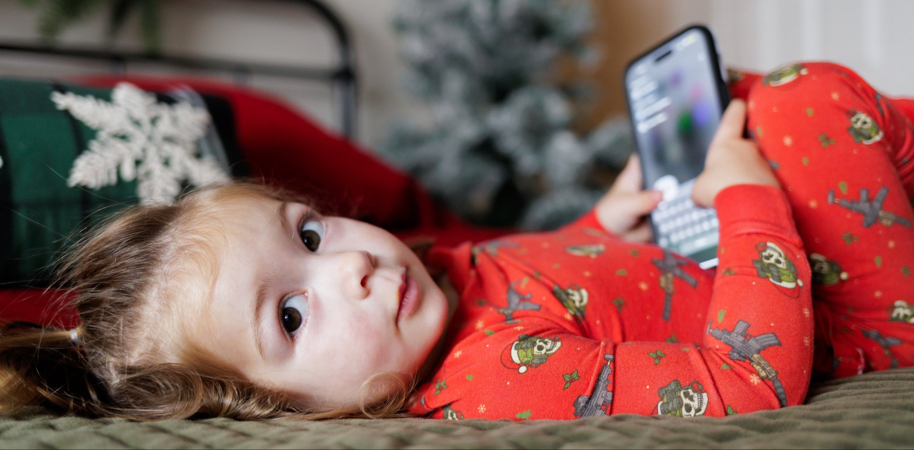 Child in red pajamas holding a smartphone on a bed with a blurred Christmas tree in the background