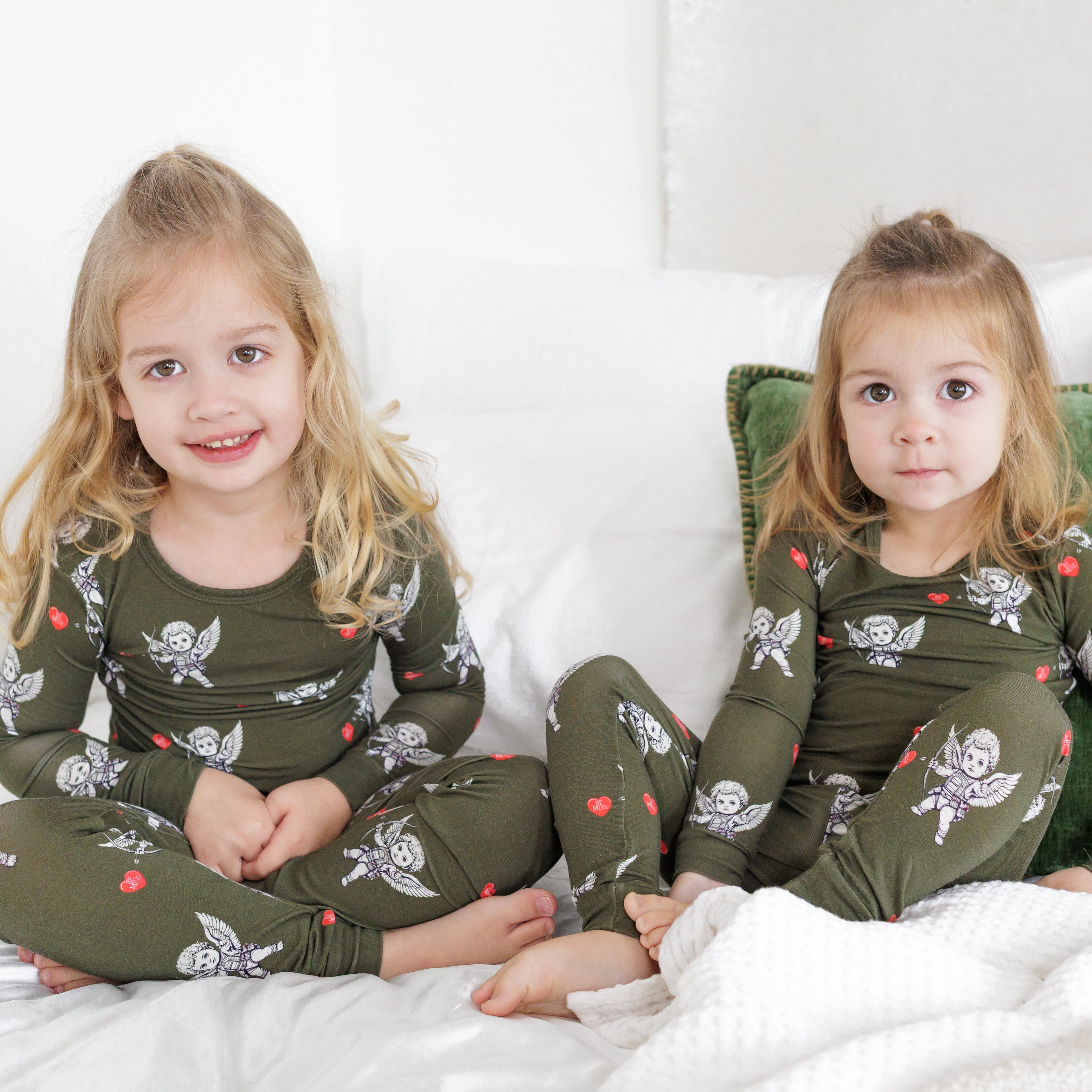 Two children in matching green pajamas sitting on a bed with white bedding and green pillows.