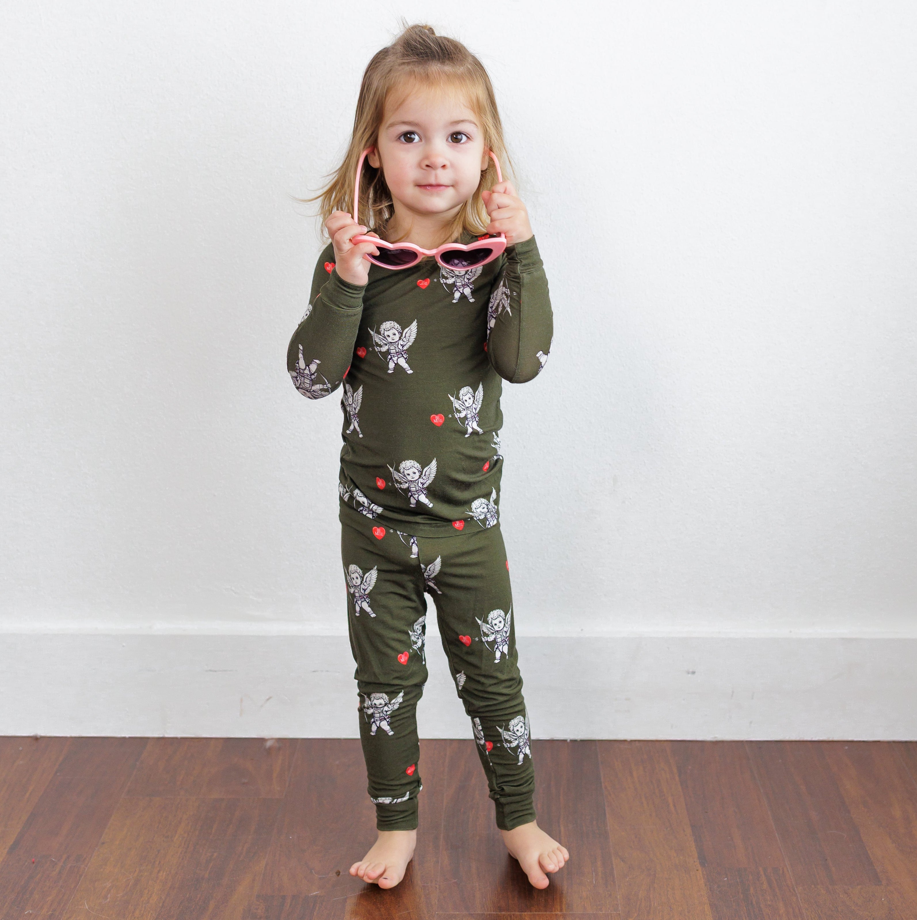 Child wearing a green pajama set with a pattern, standing on a wooden floor against a white wall.
