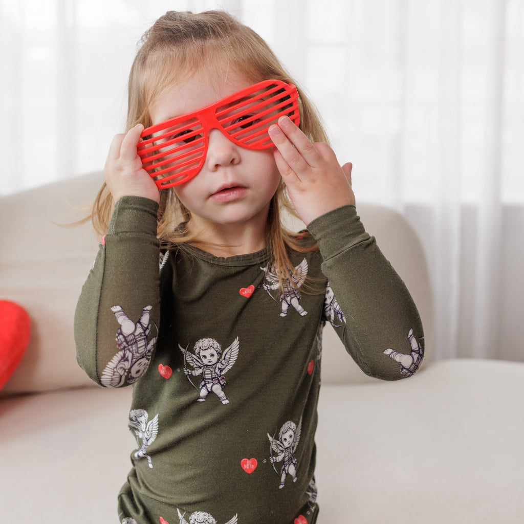Child wearing a green shirt with angel and heart designs, holding red sunglasses over their eyes.