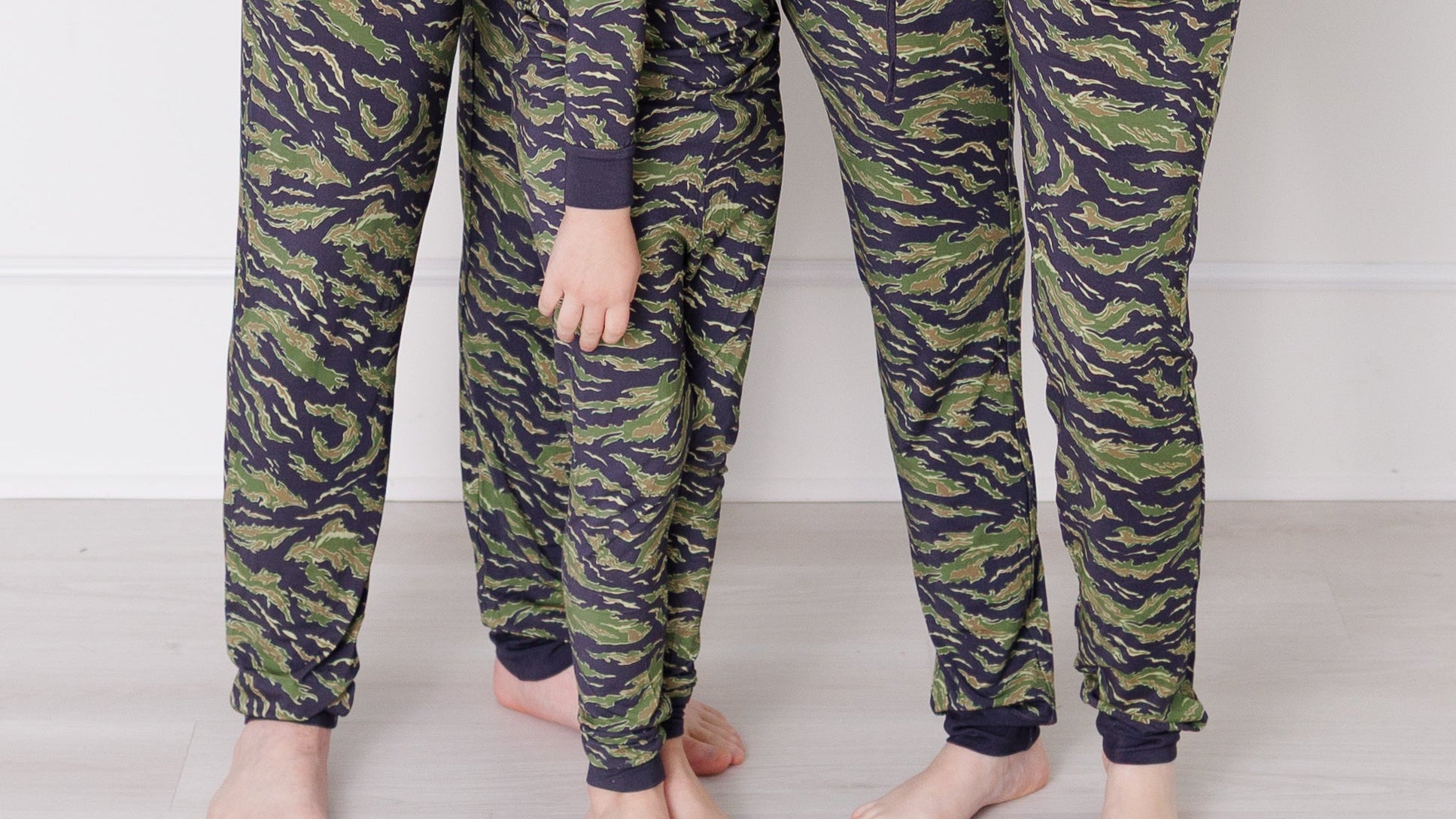 Two adults and a child wearing matching camouflage pajama pants on a plain background