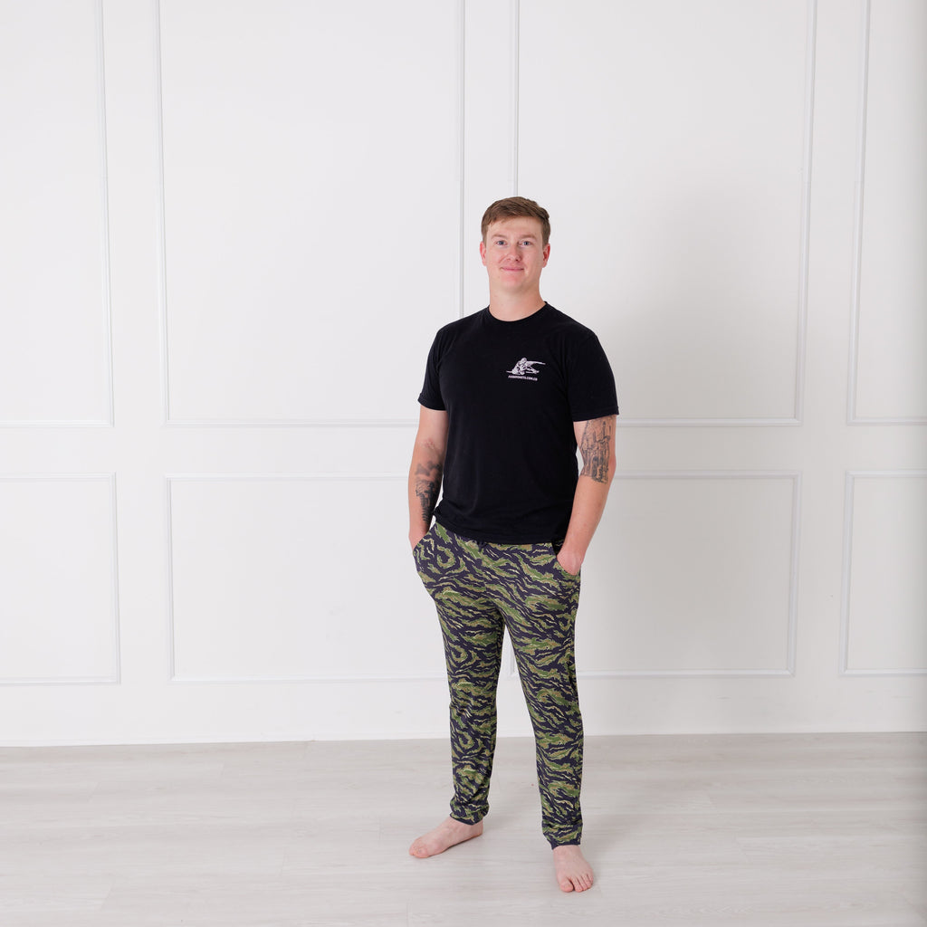 Person wearing a black t-shirt and camouflage pants standing against a white wall.