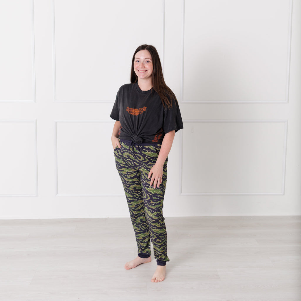 Woman wearing a black t-shirt and camouflage pants standing against a white wall.