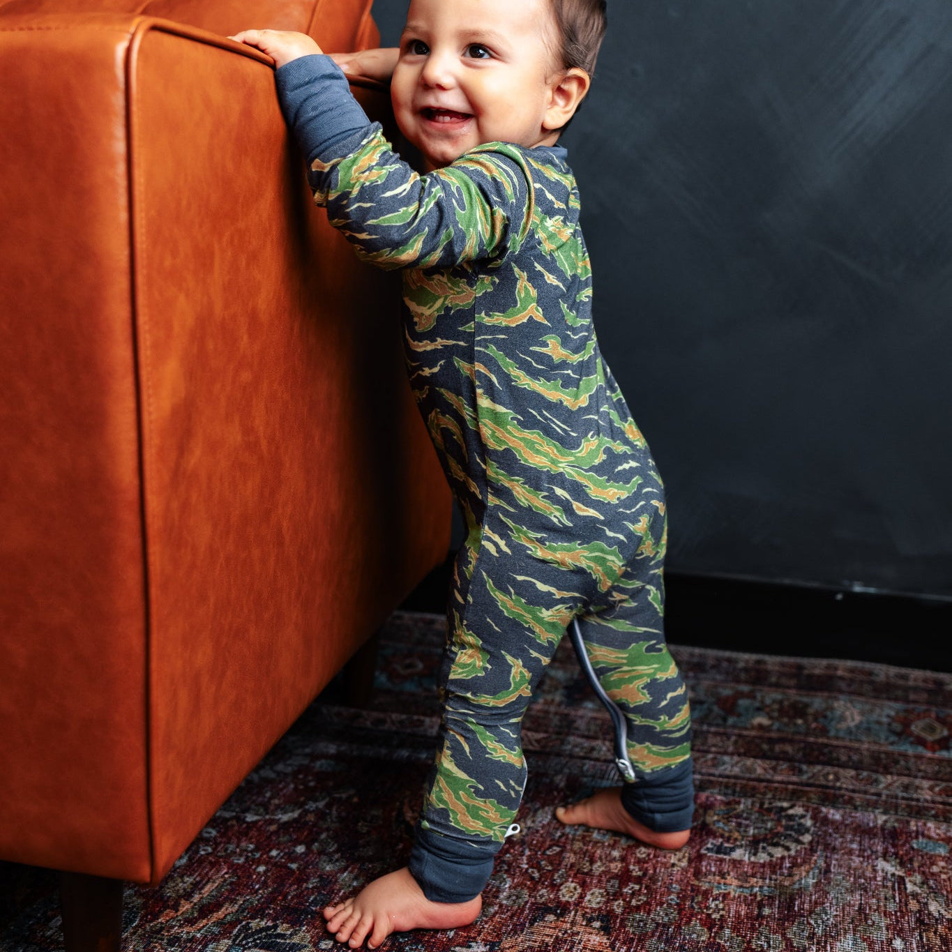 Child in a camouflage onesie standing next to an orange couch against a dark wall.
