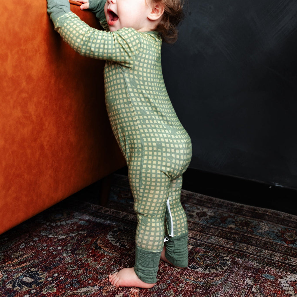 Child in green pajamas standing next to an orange couch against a dark wall.