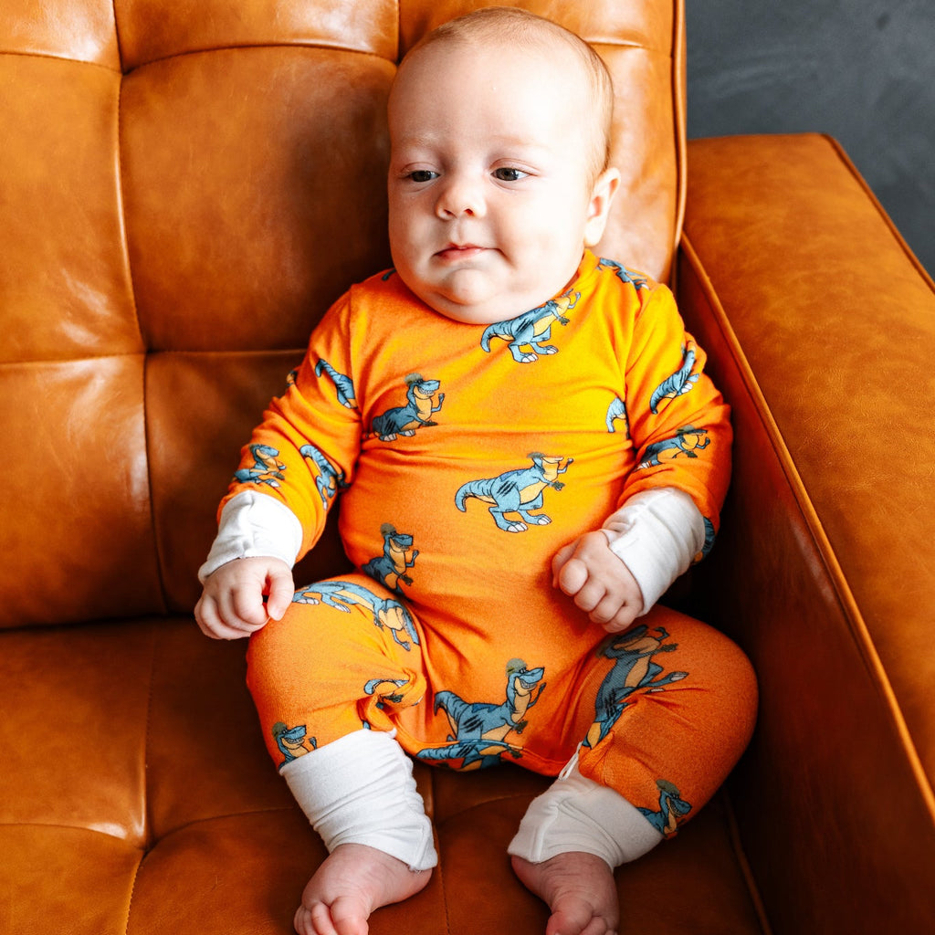 Baby in an orange outfit with dinosaur patterns sitting on a brown leather chair.