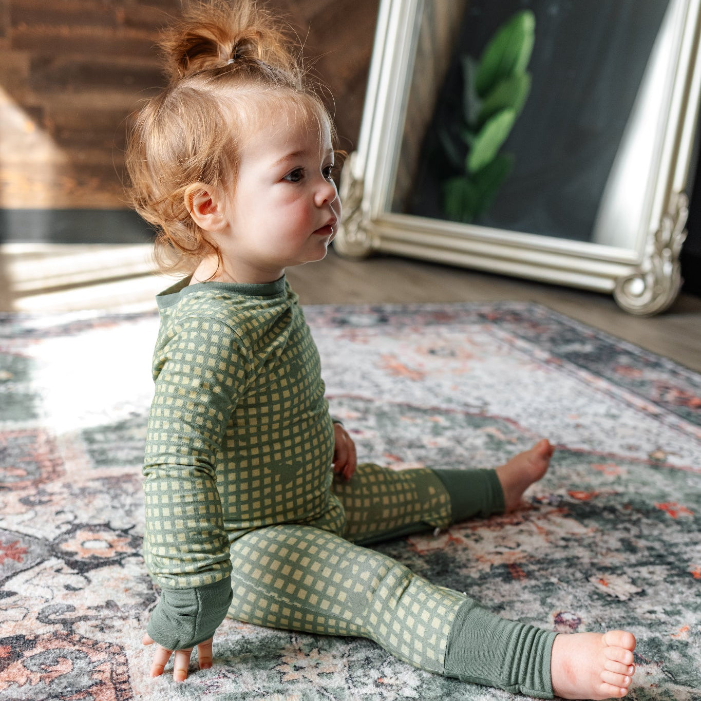 Child wearing a green patterned outfit sitting on a textured rug.