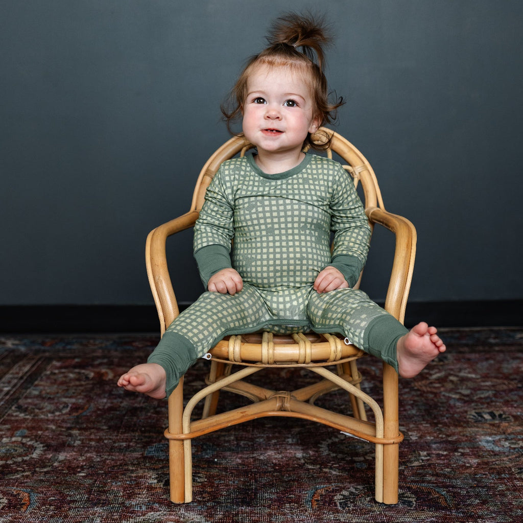 Child wearing green patterned pajamas sitting on a wooden chair against a dark gray wall.