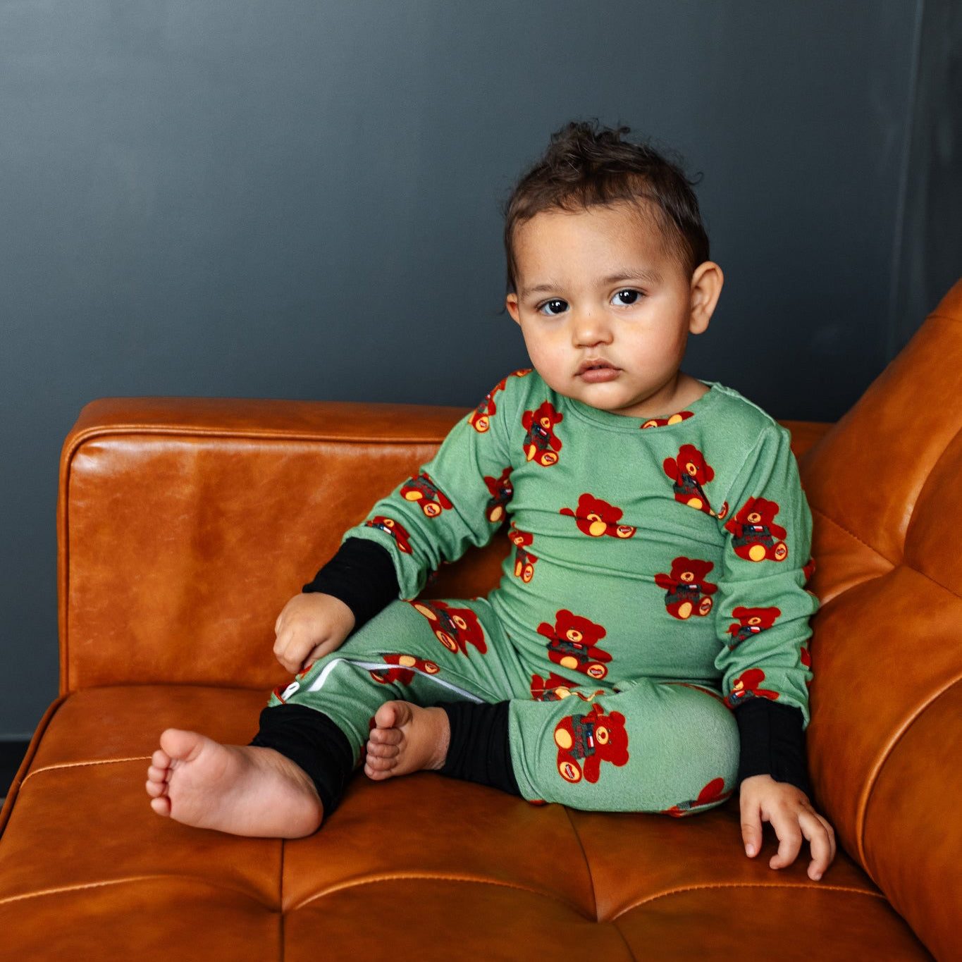 Child wearing a green outfit with red floral patterns sitting on a brown leather couch against a dark gray wall.