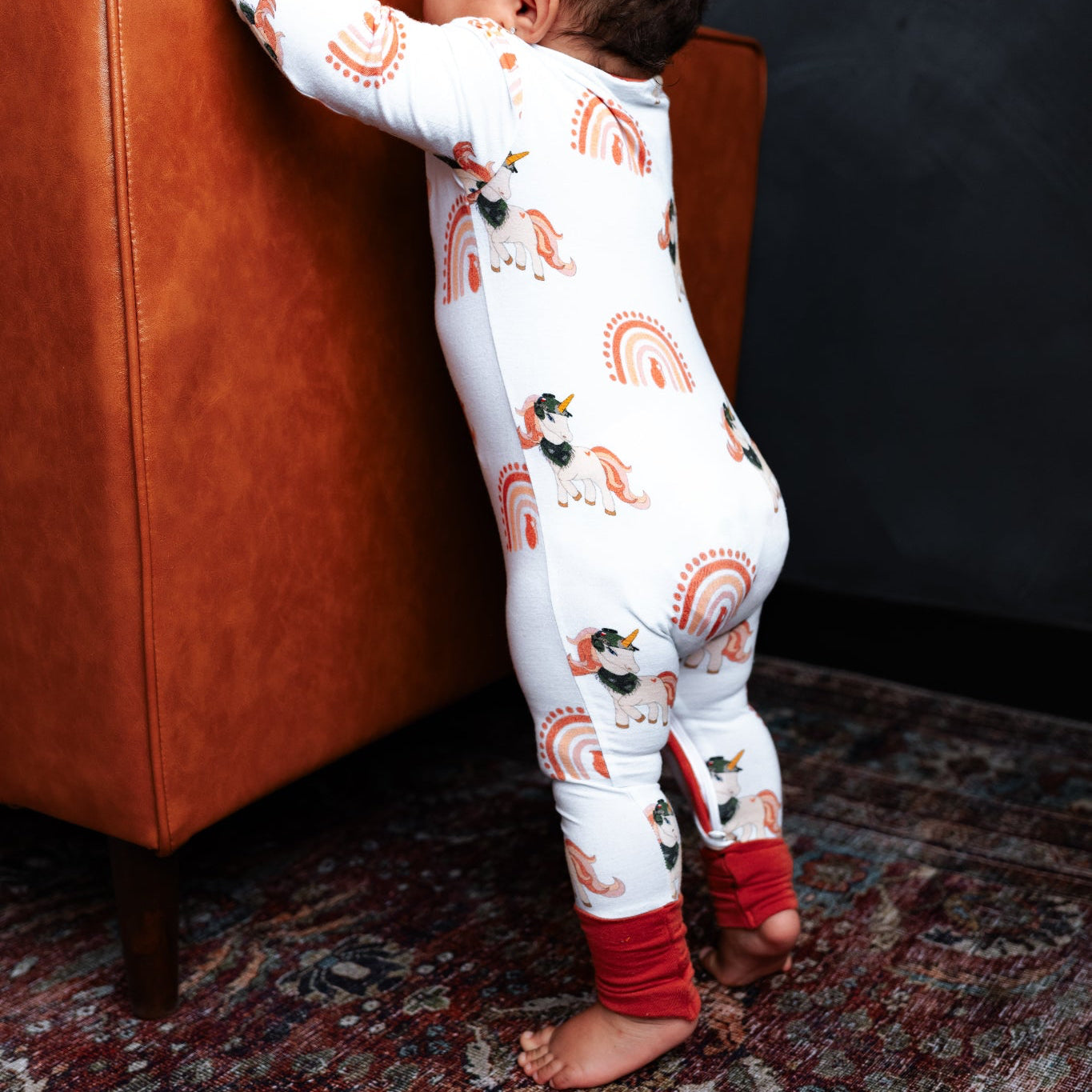 Child in a patterned onesie standing next to an orange chair on a textured rug.
