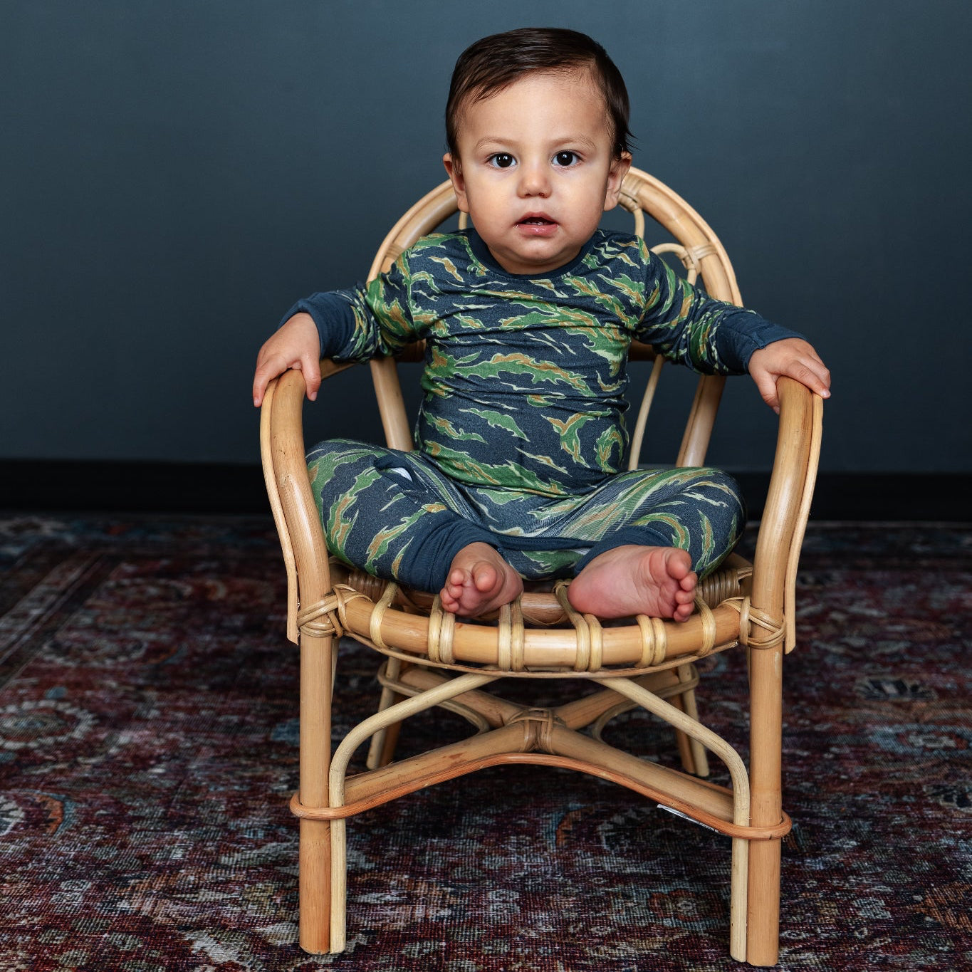 Baby in a camouflage outfit sitting on a small wooden chair against a dark background