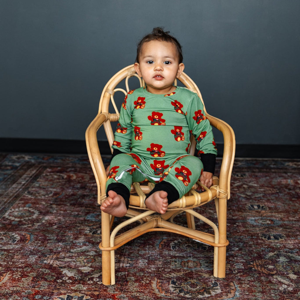 Child wearing a green floral onesie sitting on a small wooden chair against a dark gray wall.