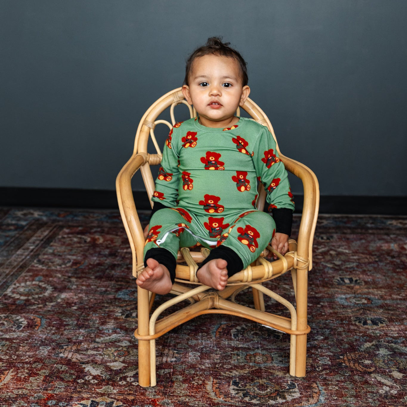 Child wearing a green floral onesie sitting on a small wooden chair against a dark gray wall.