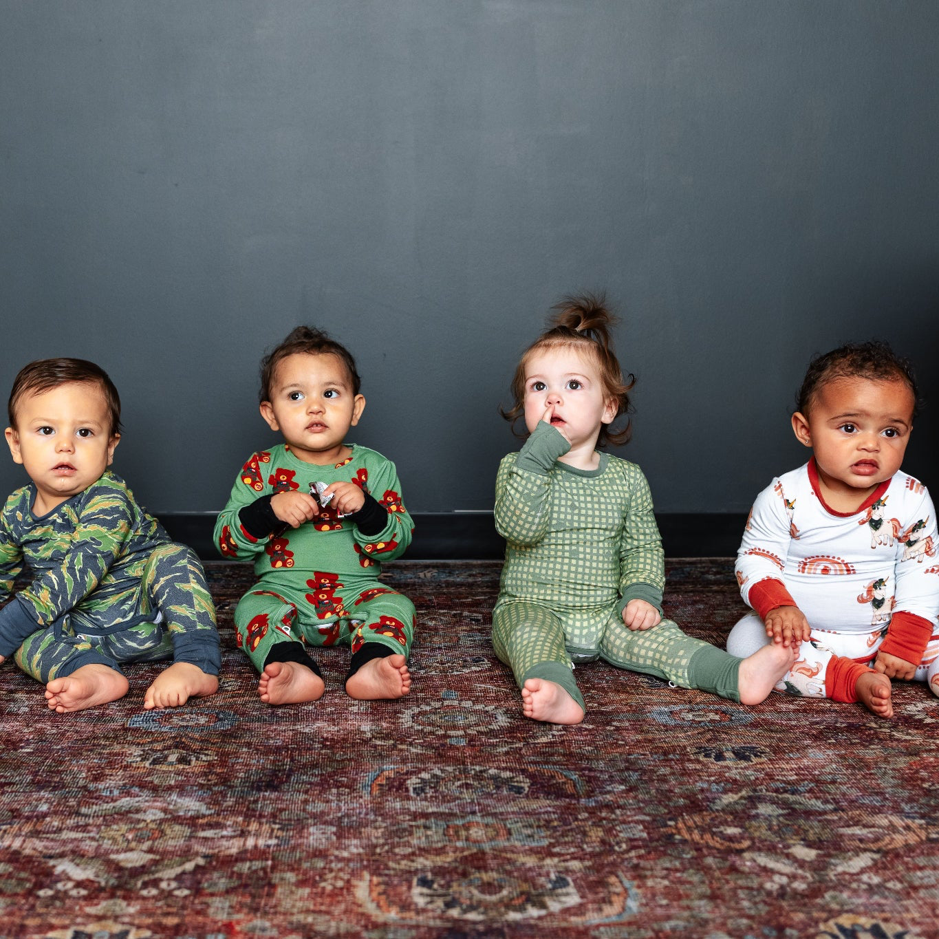 Four children in pajamas sitting on a patterned rug against a dark wall.