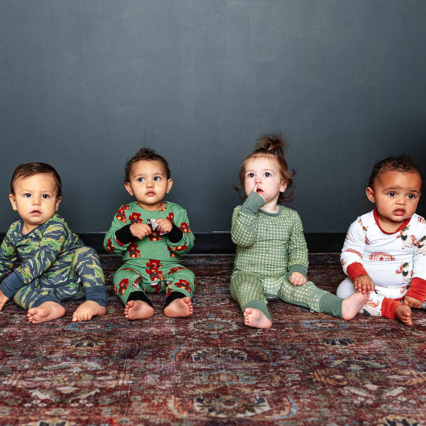 Four children in pajamas sitting on a patterned rug against a dark wall.