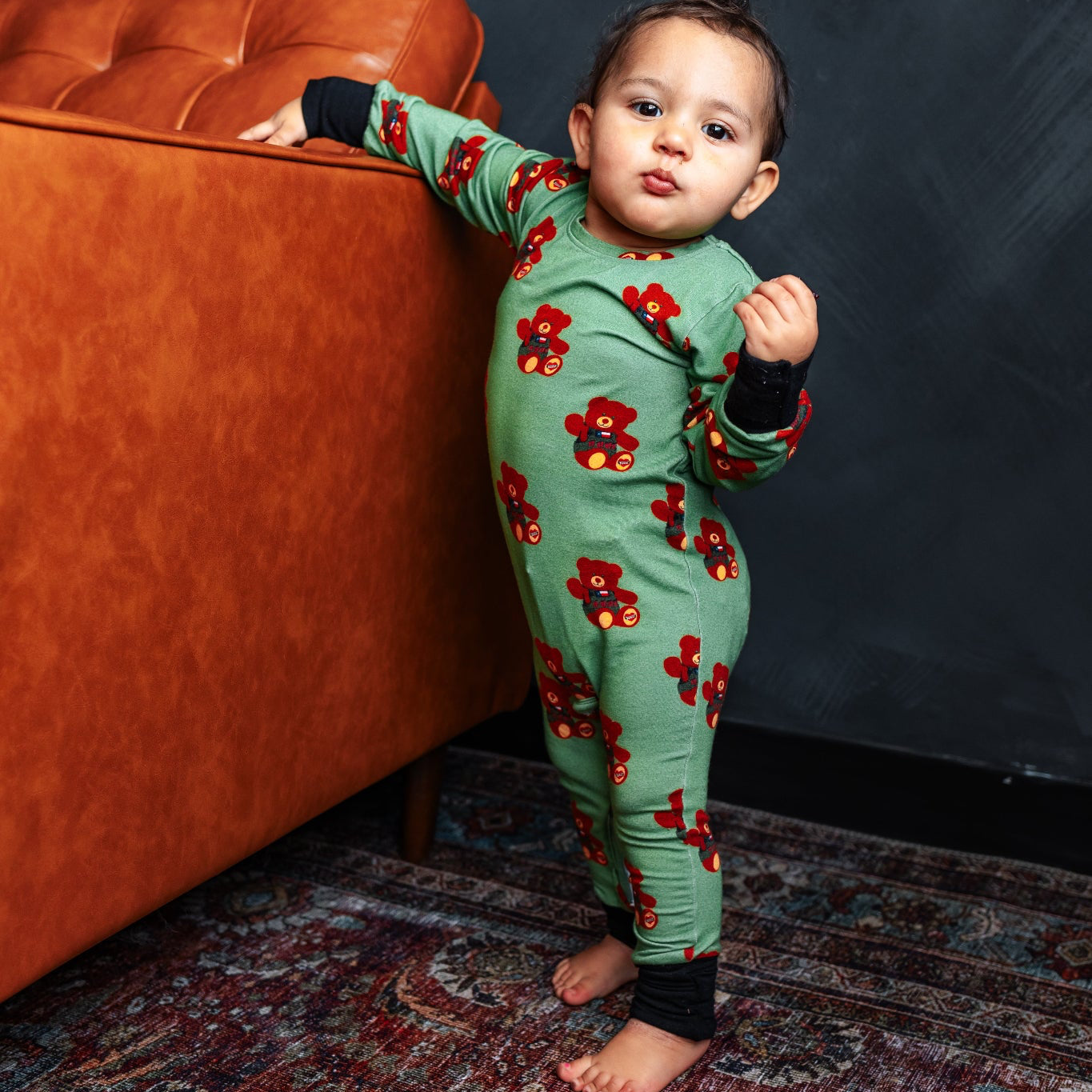 Child wearing a green onesie with red floral patterns standing next to an orange couch against a dark wall.