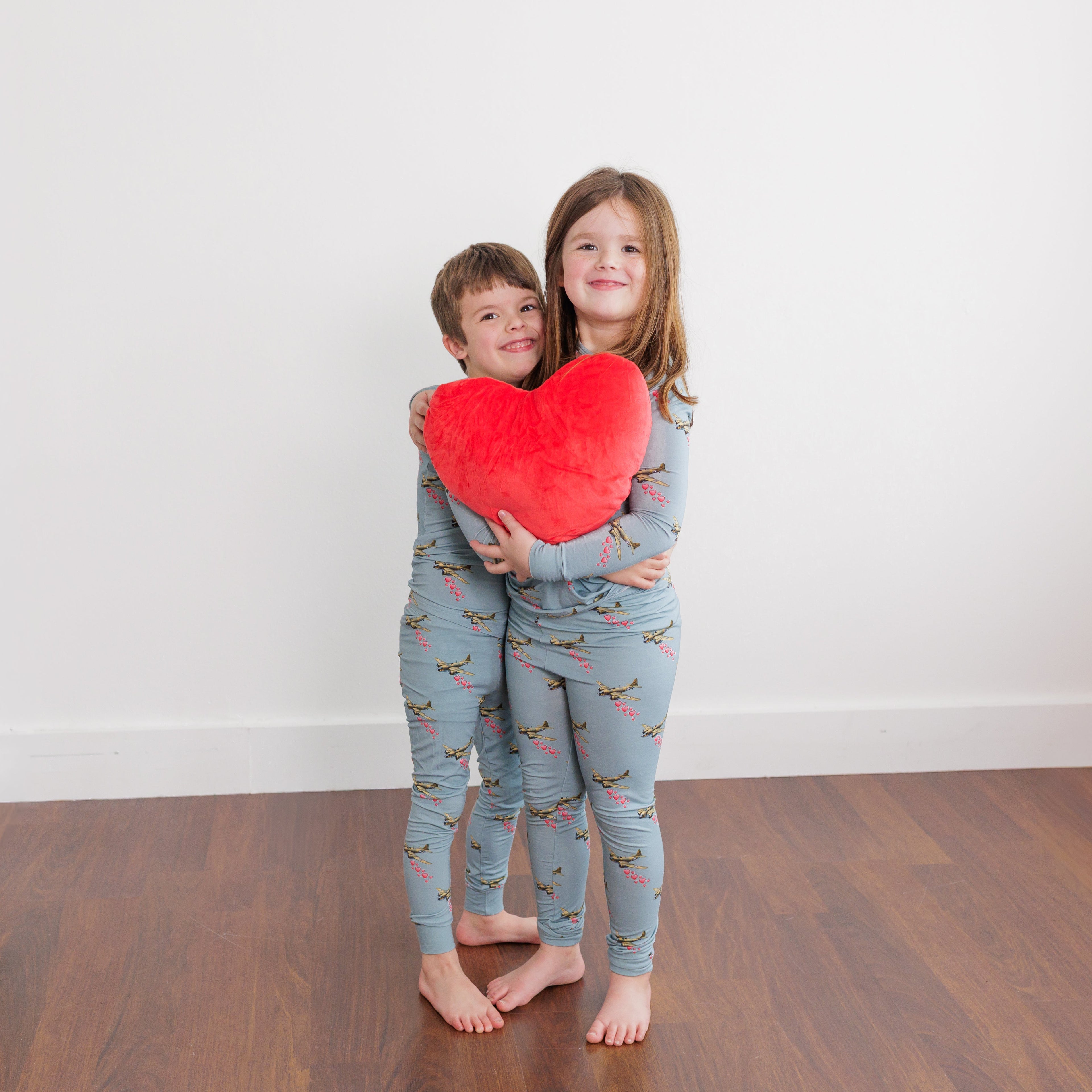 Two children holding a red heart-shaped pillow in a room with wooden flooring and white walls.