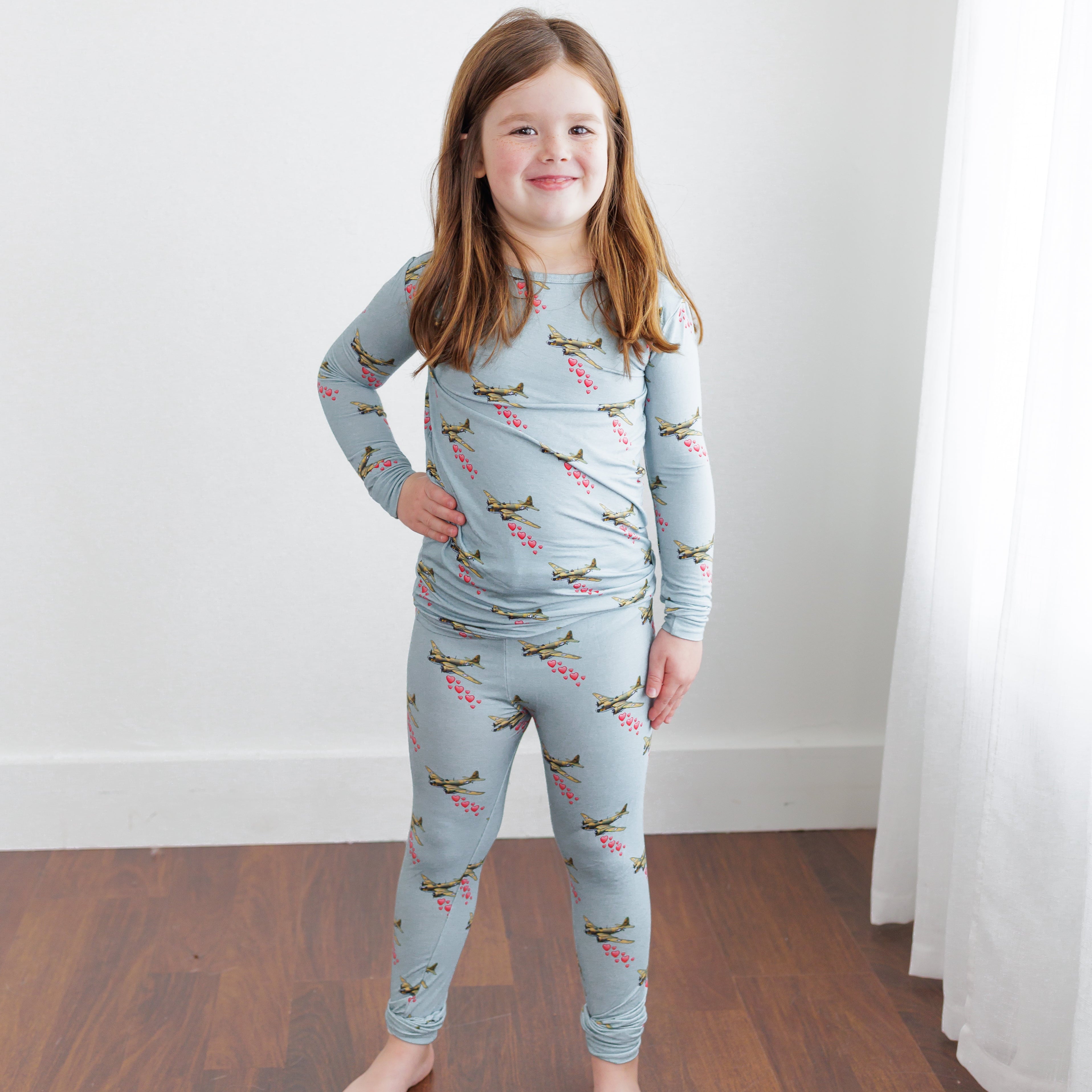 Young girl wearing a light blue pajama set with airplane pattern on a wooden floor.
